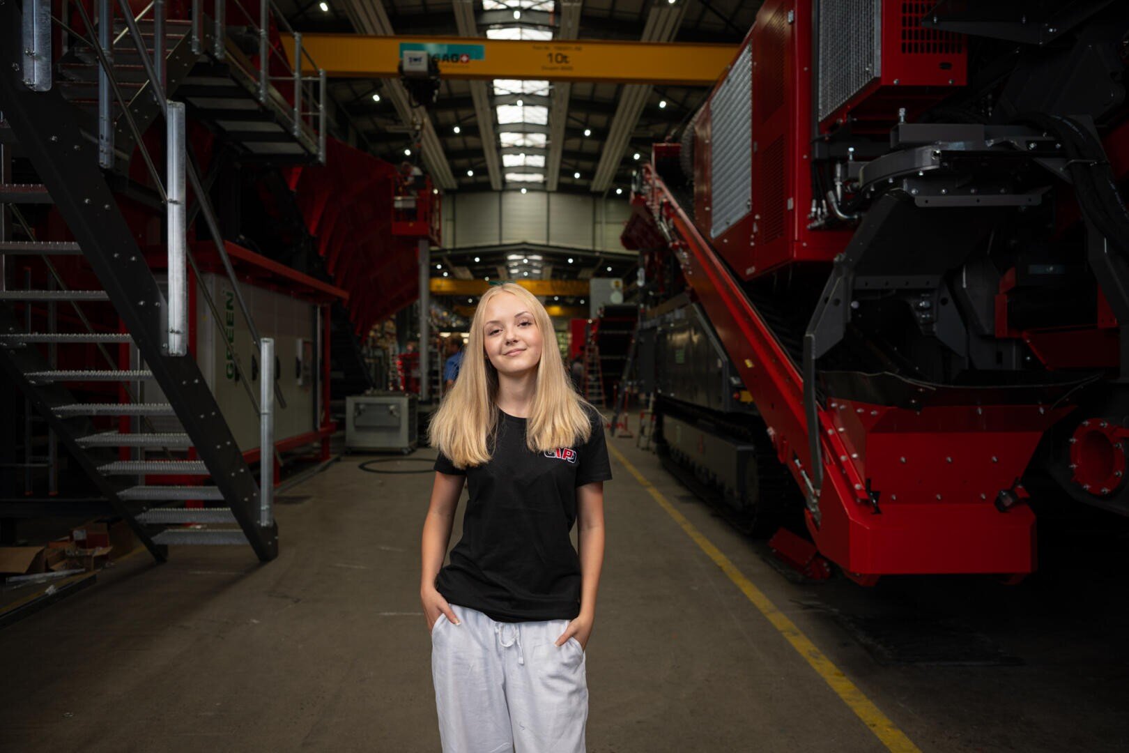 Eine junge Frau mit blonden Haaren, die ein schwarzes T-Shirt und eine weiße Hose trägt, steht lächelnd in einer großen Industriehalle, umgeben von schweren Maschinen und Geräten.