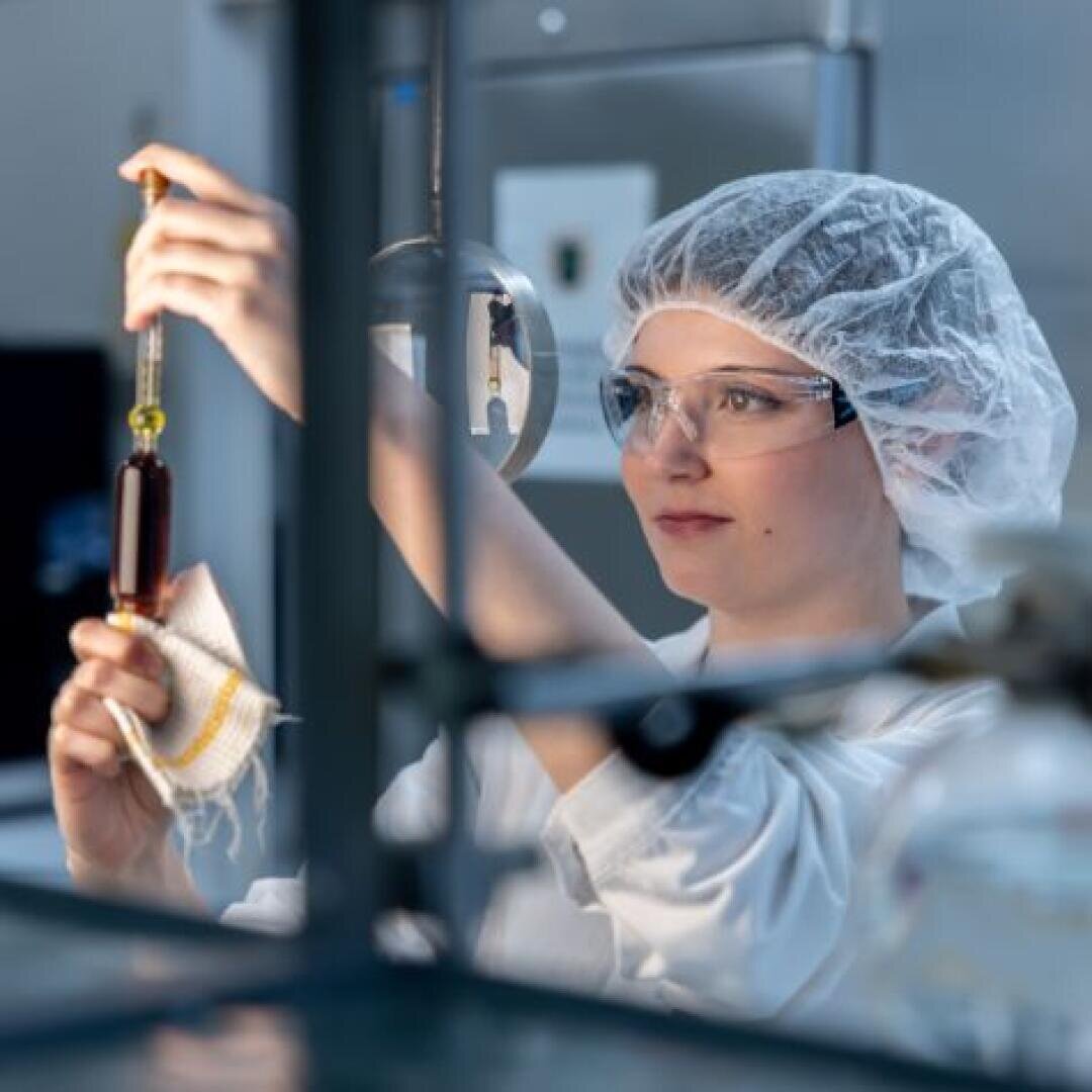 Ein Wissenschaftler mit Schutzbrille und Haarnetz überträgt in einem Labor eine dunkle Flüssigkeit mithilfe einer Pipette und konzentriert sich dabei intensiv auf den Vorgang.