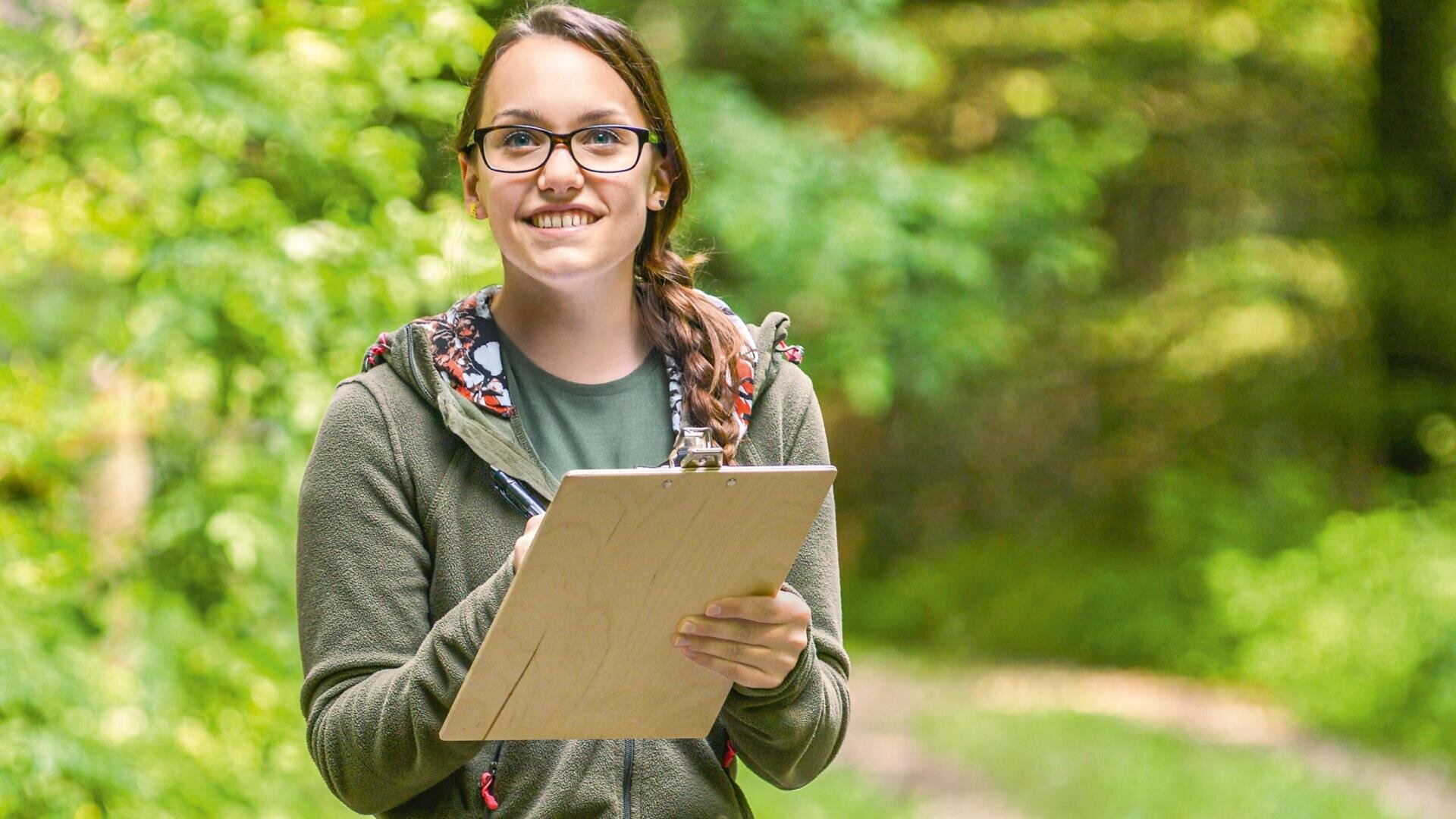 Ein lächelnder junger Mensch mit Brille und Zopf steht im Freien in einer grünen, bewaldeten Gegend und hält ein Klemmbrett und einen Stift in der Hand.