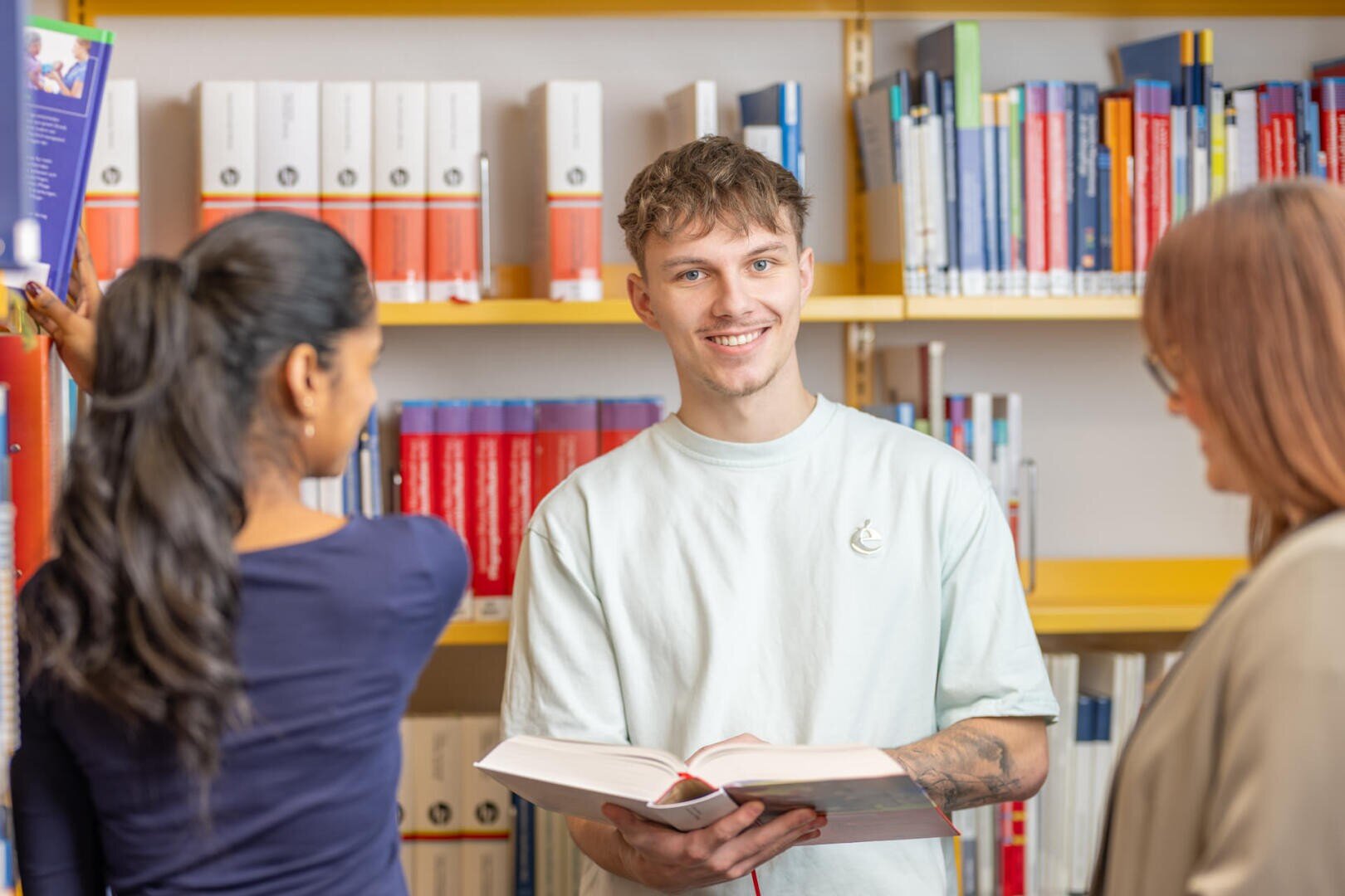 Ein junger Mann, der ein aufgeschlagenes Buch in der Hand hält, lächelt in die Kamera, während er zwischen zwei Frauen vor den mit bunten Büchern gefüllten Bücherregalen einer Bibliothek steht.