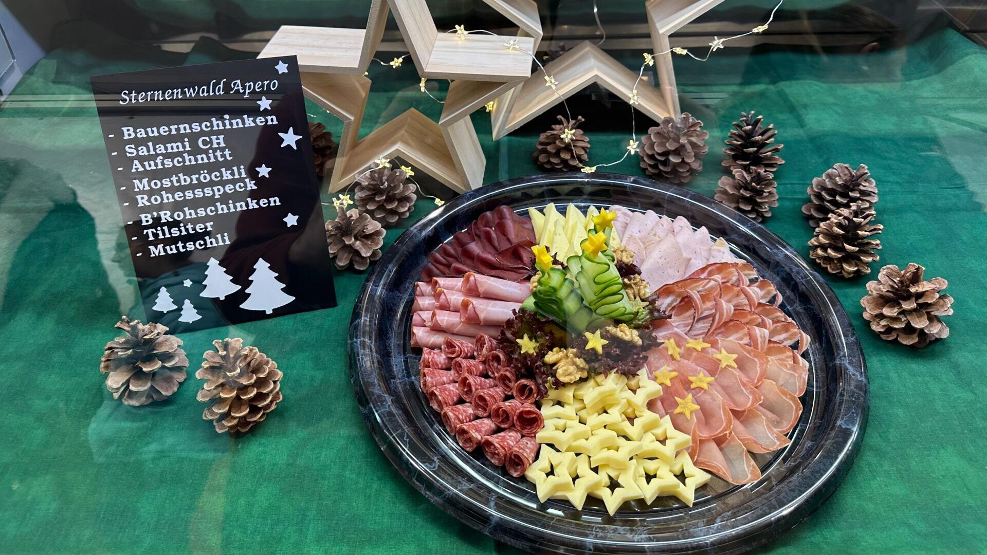 Auf einer grünen Tischdecke steht eine Platte mit verschiedenen Aufschnitt-Sorten, sternförmig geschnittenem Käse und Beilagen. Daneben sind Tannenzapfen, Holzsterne und ein Schild mit einer Auflistung der Fleisch- und Käsesorten ausgestellt.