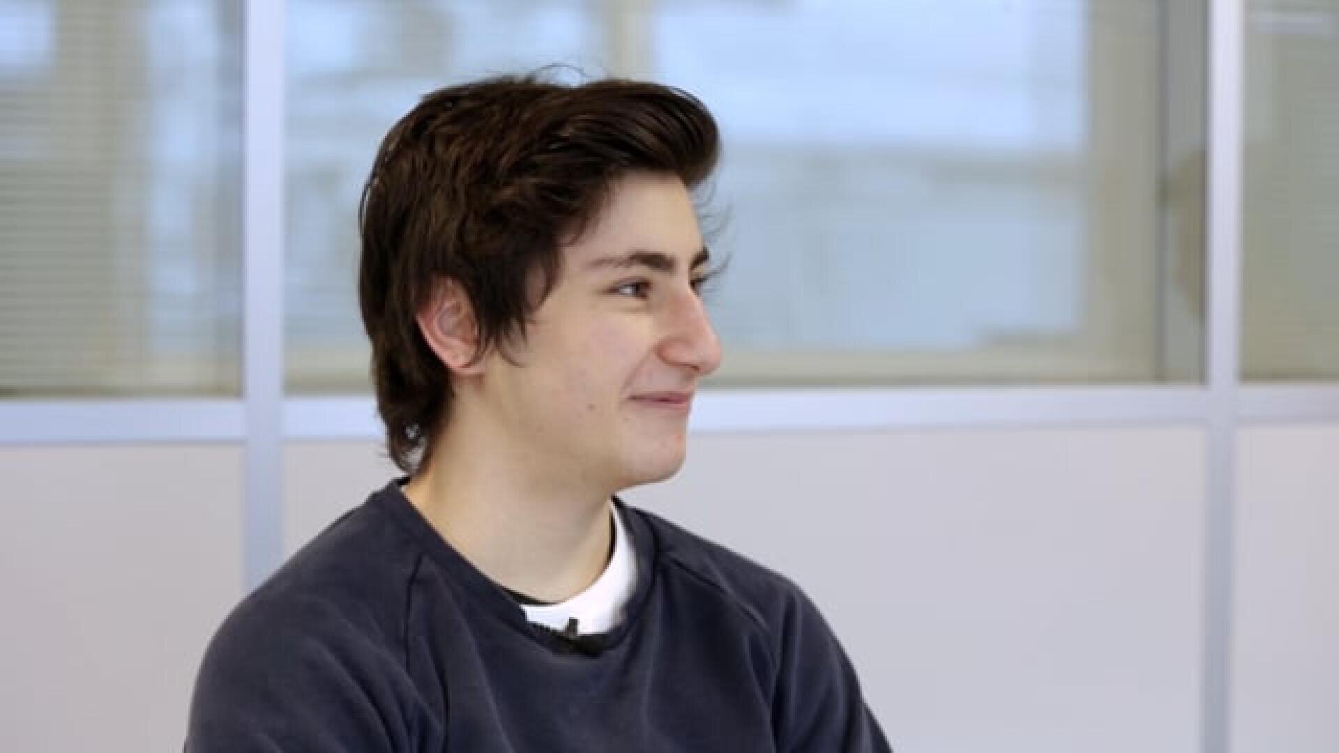 Eine junge Person mit kurzen dunklen Haaren, die ein dunkles Sweatshirt trägt, sitzt in einem Haus und lächelt, während sie nach links schaut. Der Hintergrund ist unscharf und zeigt Fenster und helle Wände.