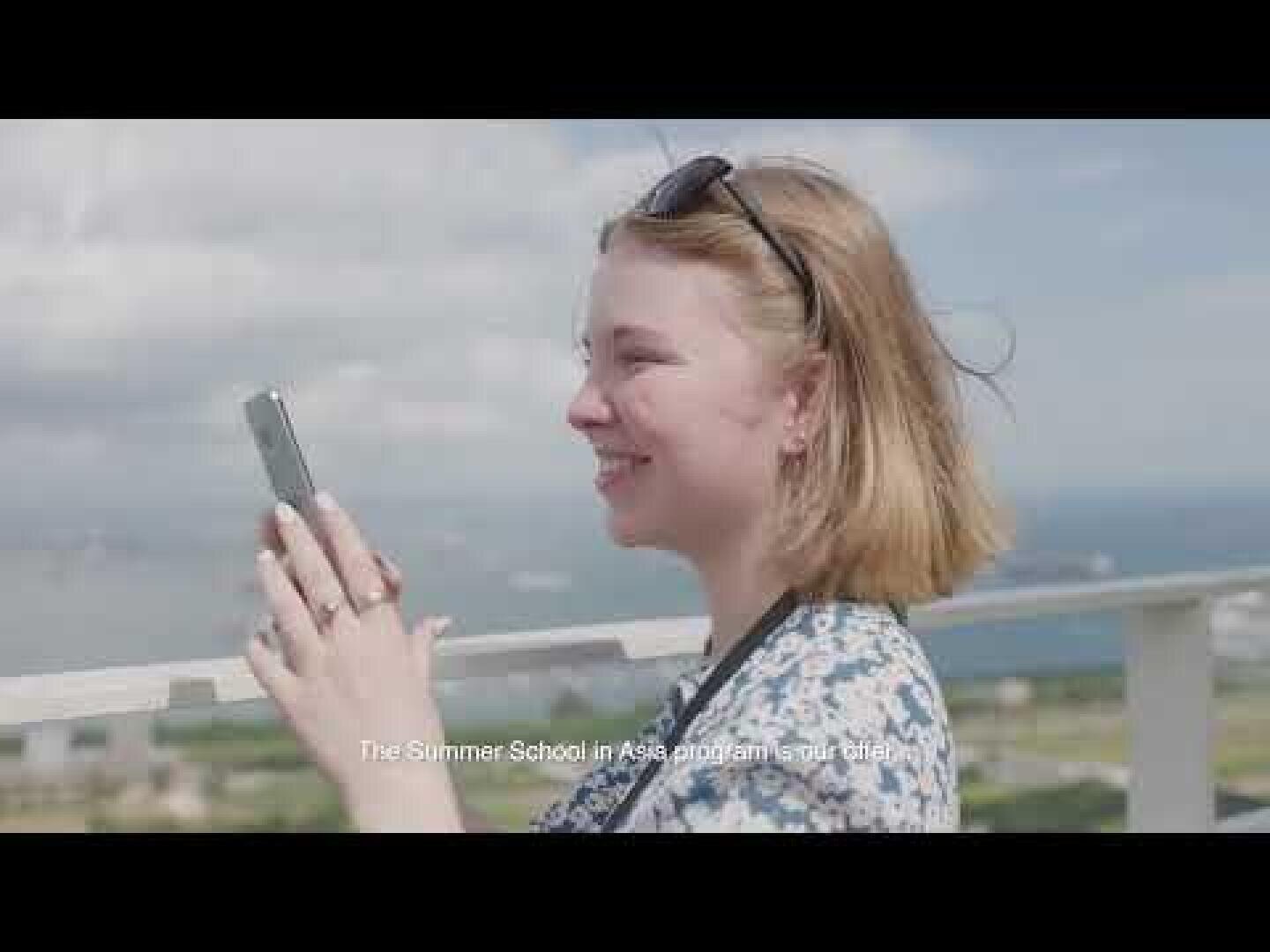 Eine junge Frau mit kurzen blonden Haaren lächelt, während sie im Freien mit ihrem Smartphone ein Foto macht – im Hintergrund sind das Meer und Schiffe zu sehen. Im Untertitel unten steht: „Das Programm ‚Sommerschule in Asien‘ hat begonnen.“.