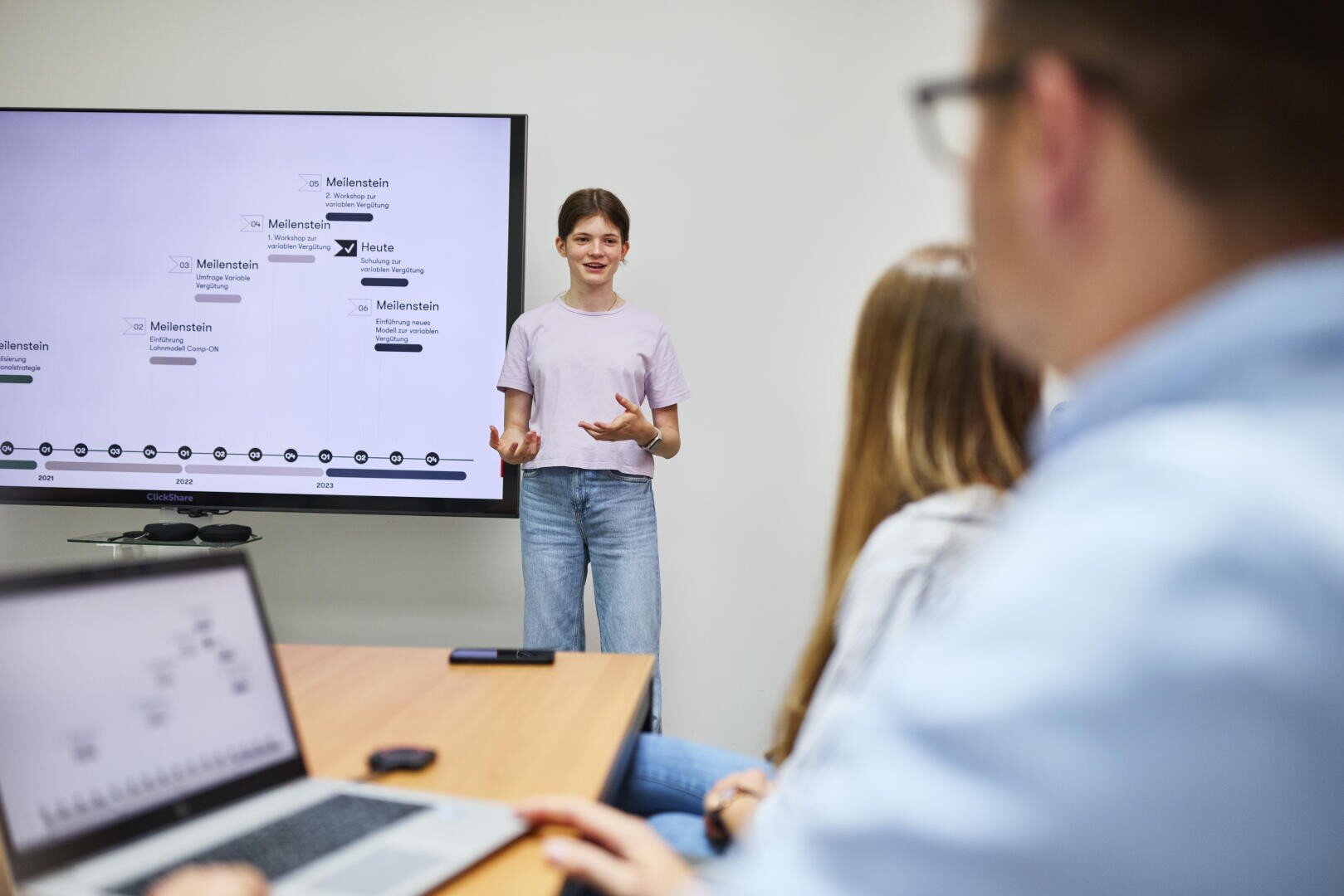 Eine junge Frau steht vor einem großen Bildschirm, auf dem ein Zeitdiagramm zu sehen ist, während zwei Personen mit Laptops an einem Tisch sitzen und in einem Besprechungsraum aufmerksam zuhören.