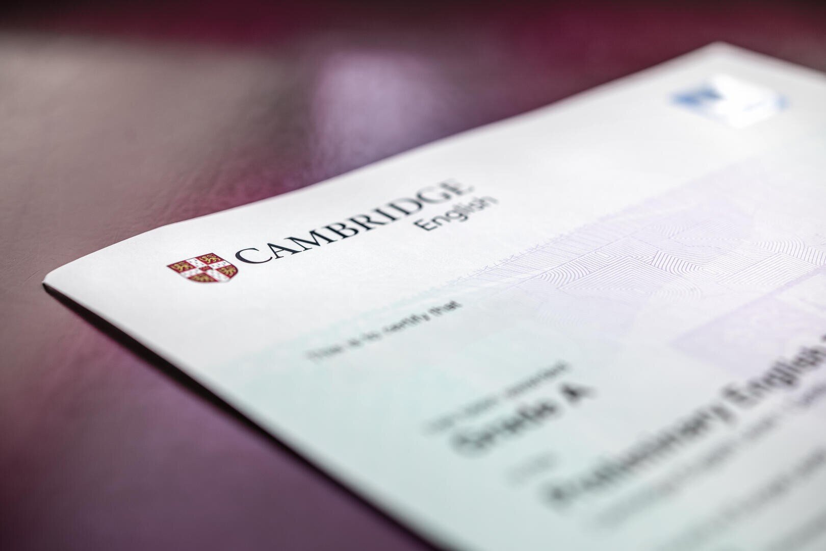 Nahaufnahme eines Cambridge-Englisch-Zertifikats auf einer kastanienbraunen Oberfläche, die das Cambridge-Logo und einen Teil des Textes zeigt, wobei das Zertifikat an den Rändern leicht unscharf ist.