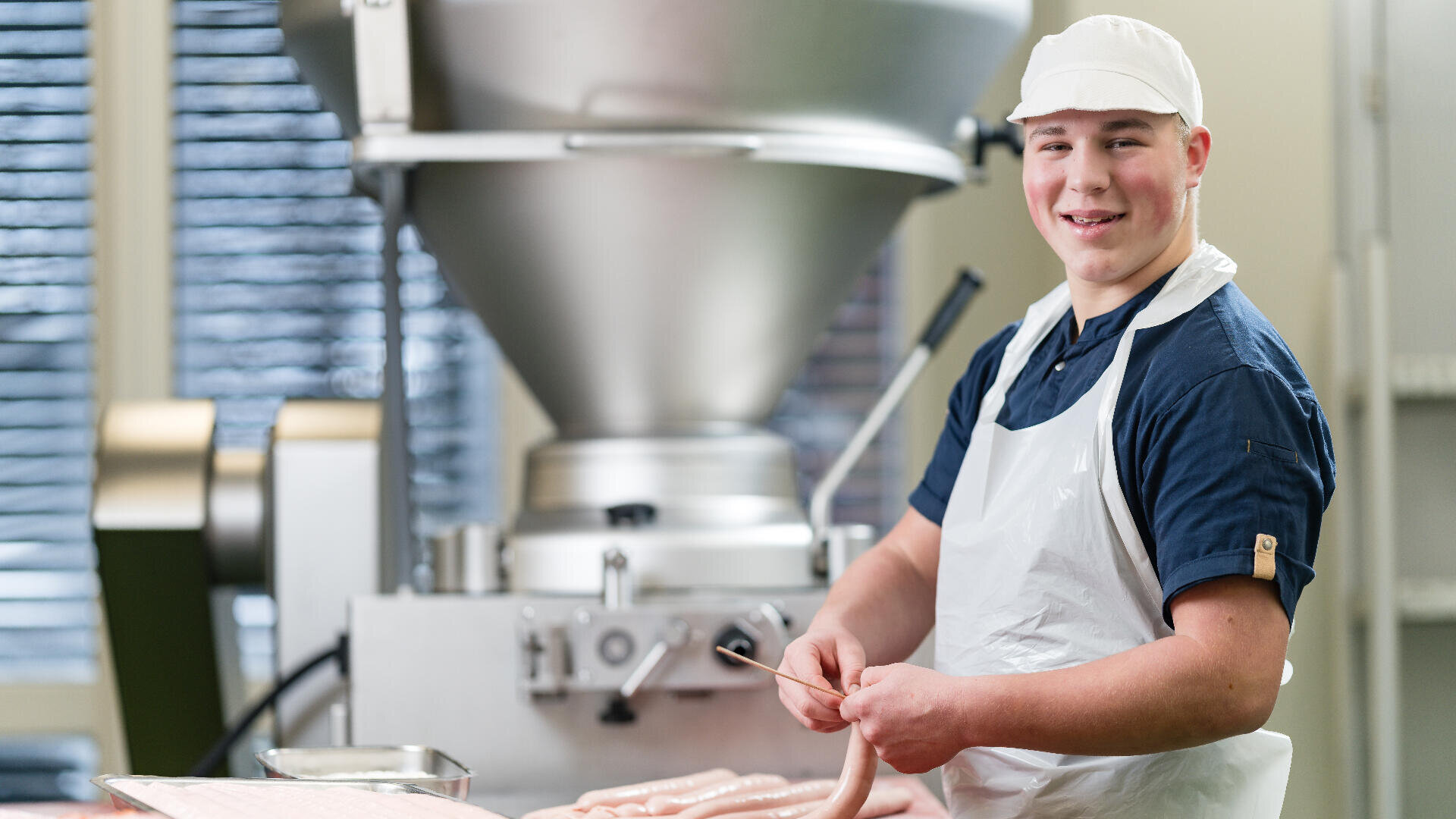 Ein lächelnder Metzger mit weißer Schürze und Mütze steht vor einer industriellen Wurstherstellungsanlage, hält eine Wursthülle in der Hand und bereitet Fleischprodukte in einer sauberen Lebensmittelverarbeitungsanlage zu.