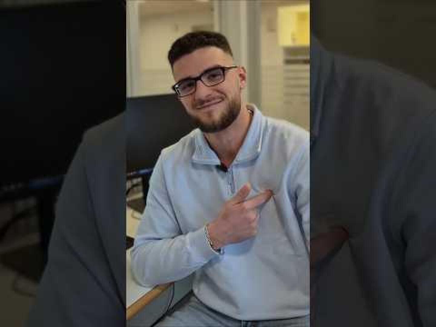 Ein Mann mit kurzen dunklen Haaren, Brille und Bart sitzt in einem Raum, lächelt und zeigt mit einer Hand nach links. Er trägt einen hellgrauen Pullover mit Reißverschluss.
