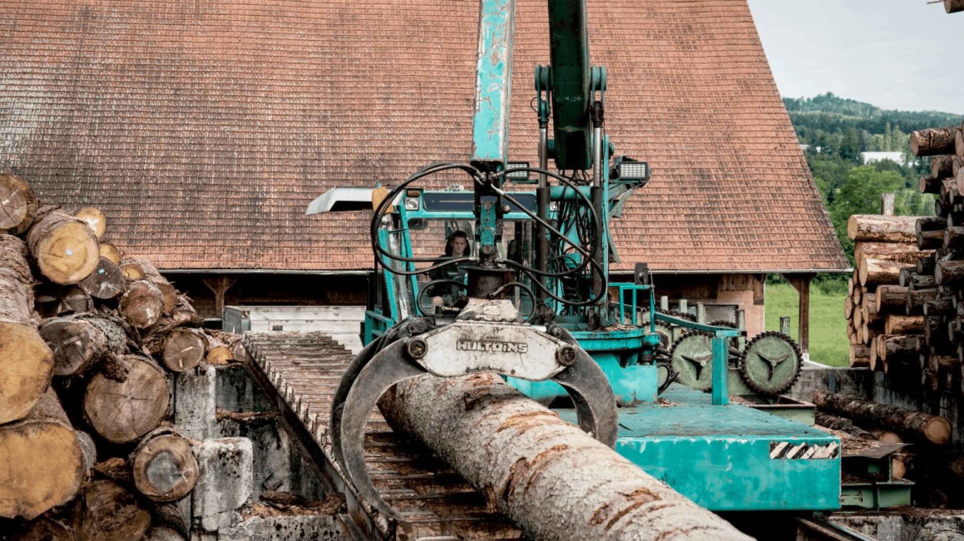 Eine blaugrüne industrielle Holzerntemaschine greift und hebt einen großen Baumstamm in einem Holzlager, umgeben von gestapelten Stämmen und einem Gebäude mit rotem Ziegeldach im Hintergrund.