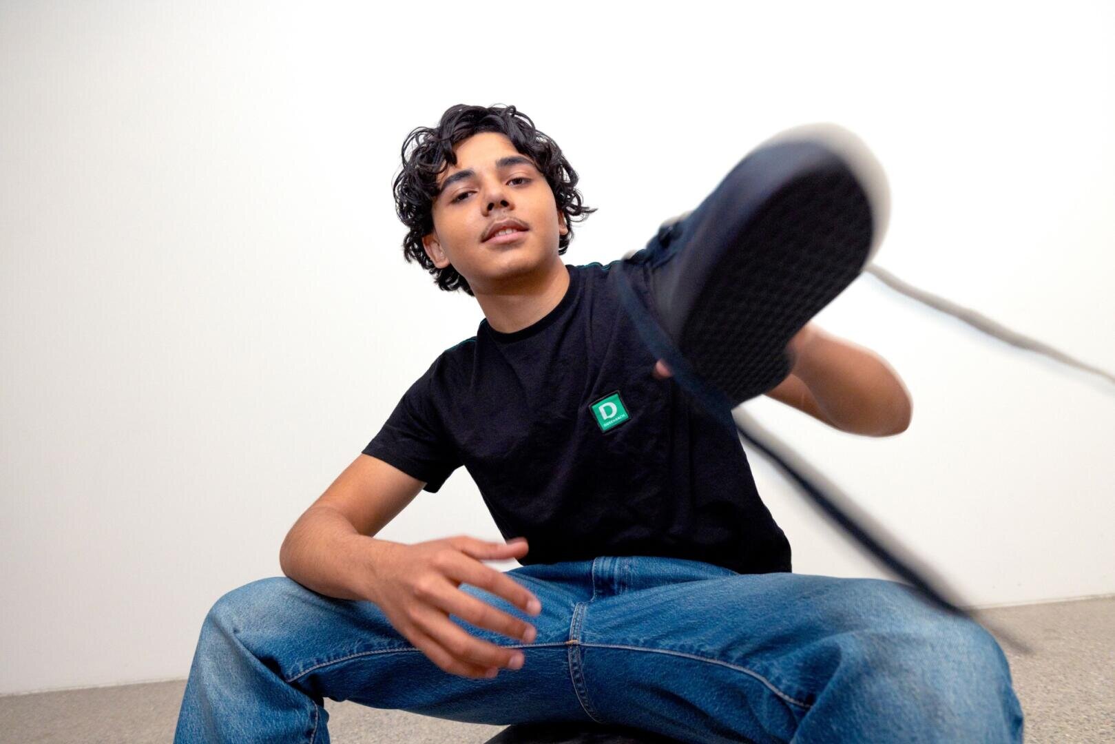 Eine junge Person in schwarzem T-Shirt und blauer Jeans sitzt auf einem Hocker vor einer weißen Wand und hebt ein Bein nach vorne, wobei der Fuß nahe an der Kamera steht, wodurch eine dynamische Perspektive entsteht.