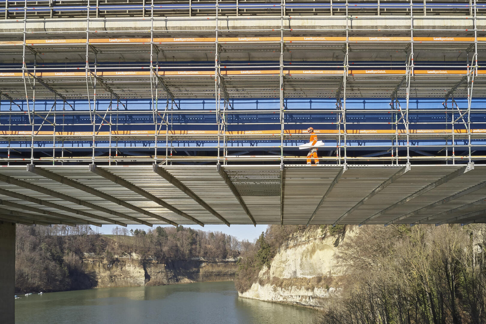 Ein Bauarbeiter in orangefarbener Schutzkleidung geht auf einem Gerüst unter einer großen Brücke über einen Fluss entlang, im Hintergrund sind Felsen und Bäume zu sehen.
