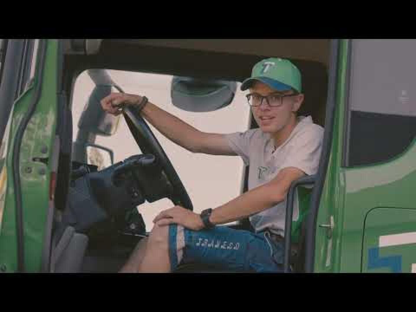 Ein junger Mann mit Brille, grüner Mütze und T-Shirt sitzt auf dem Fahrersitz eines grünen Lieferwagens und lächelt in die Kamera.