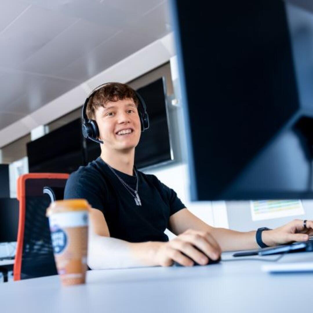 Ein junger Mensch mit einem Headset lächelt, während er in einem modernen Büro einen Computer benutzt. Ein Kaffeebecher zum Mitnehmen steht auf dem Schreibtisch neben ihr.