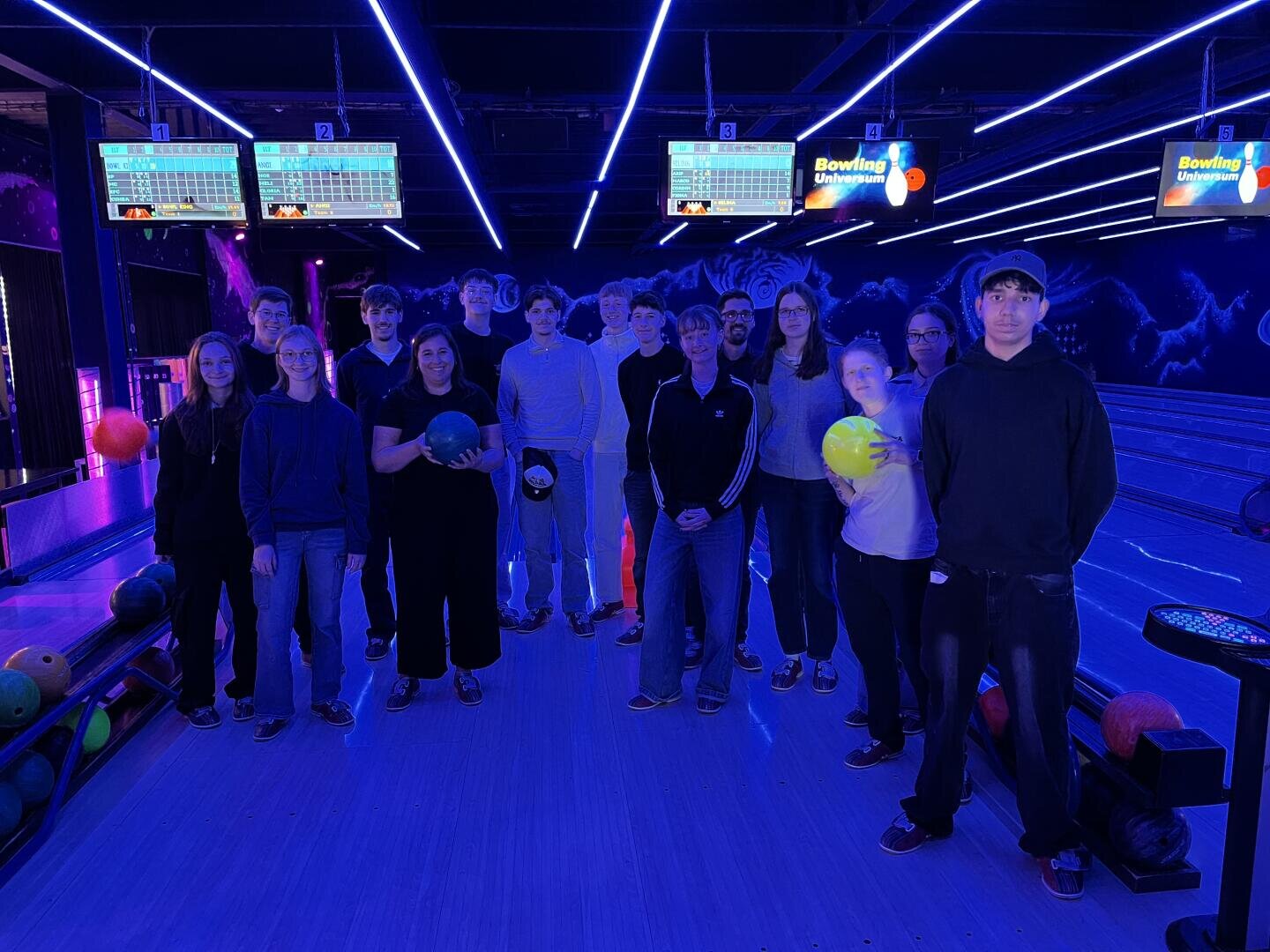 Eine Gruppe von Menschen steht in einer neonbeleuchteten Bowlingbahn, einige halten Bowlingkugeln in den Händen; über ihnen hängen Anzeigetafeln, und bunte Lichter tauchen die Szene in ein helles Licht.