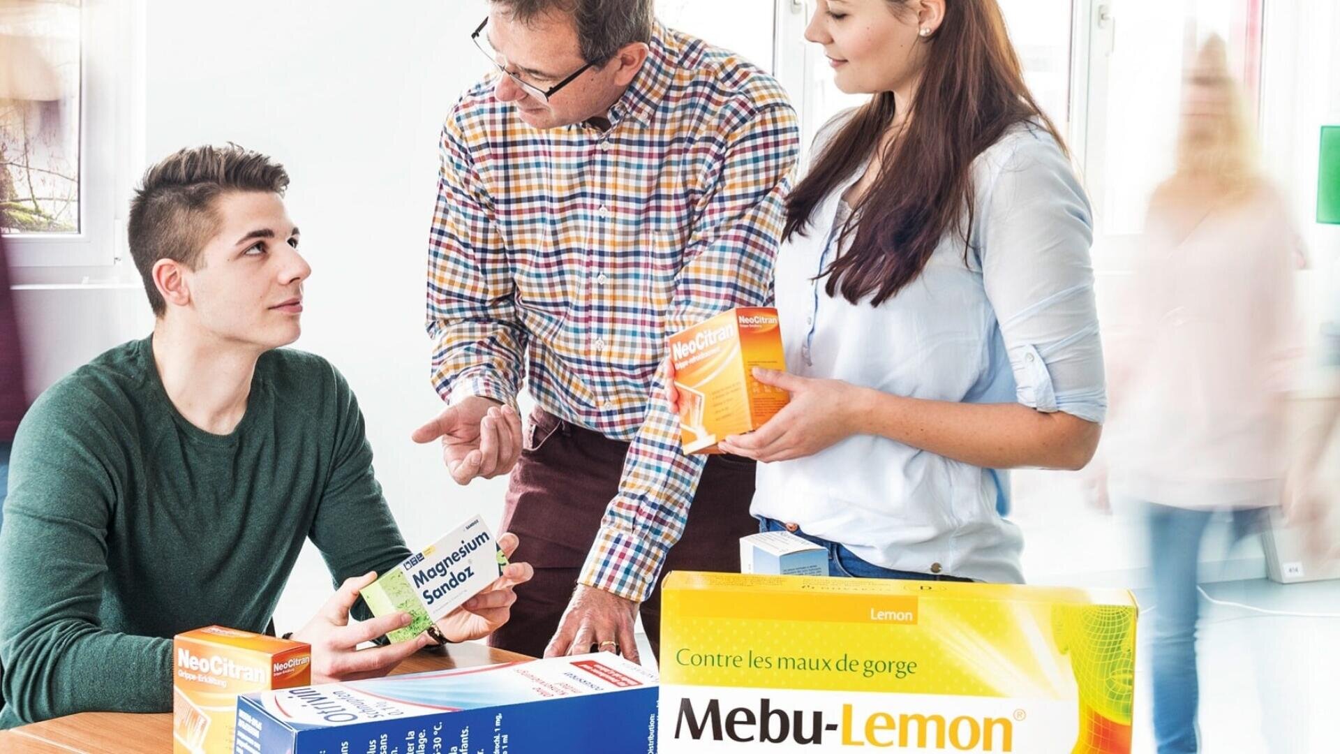Drei Personen diskutieren in einem hellen Raum über rezeptfreie Medikamente und halten Schachteln mit der Aufschrift Mebu-Lemon und Monapure Sandoz in der Hand. Große Medikamentenpackungen liegen auf dem Tisch vor ihnen. Zwei verschwommene Gestalten laufen im Hintergrund.