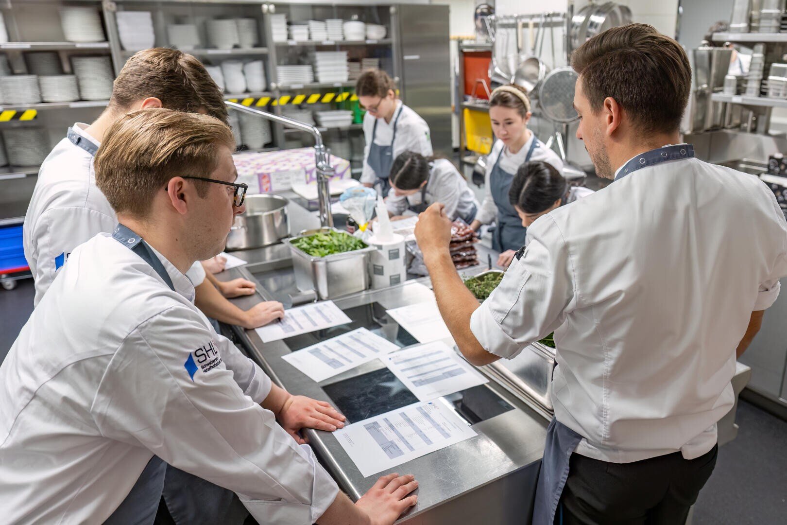 Eine Gruppe von Köchen in weißen Uniformen versammelt sich um eine Arbeitsplatte aus rostfreiem Stahl in einer Großküche, um gedruckte Speisekarten oder Rezepte zu prüfen und Vorbereitungen zu besprechen. Im Hintergrund sind verschiedene Küchengeräte und Zutaten zu sehen.