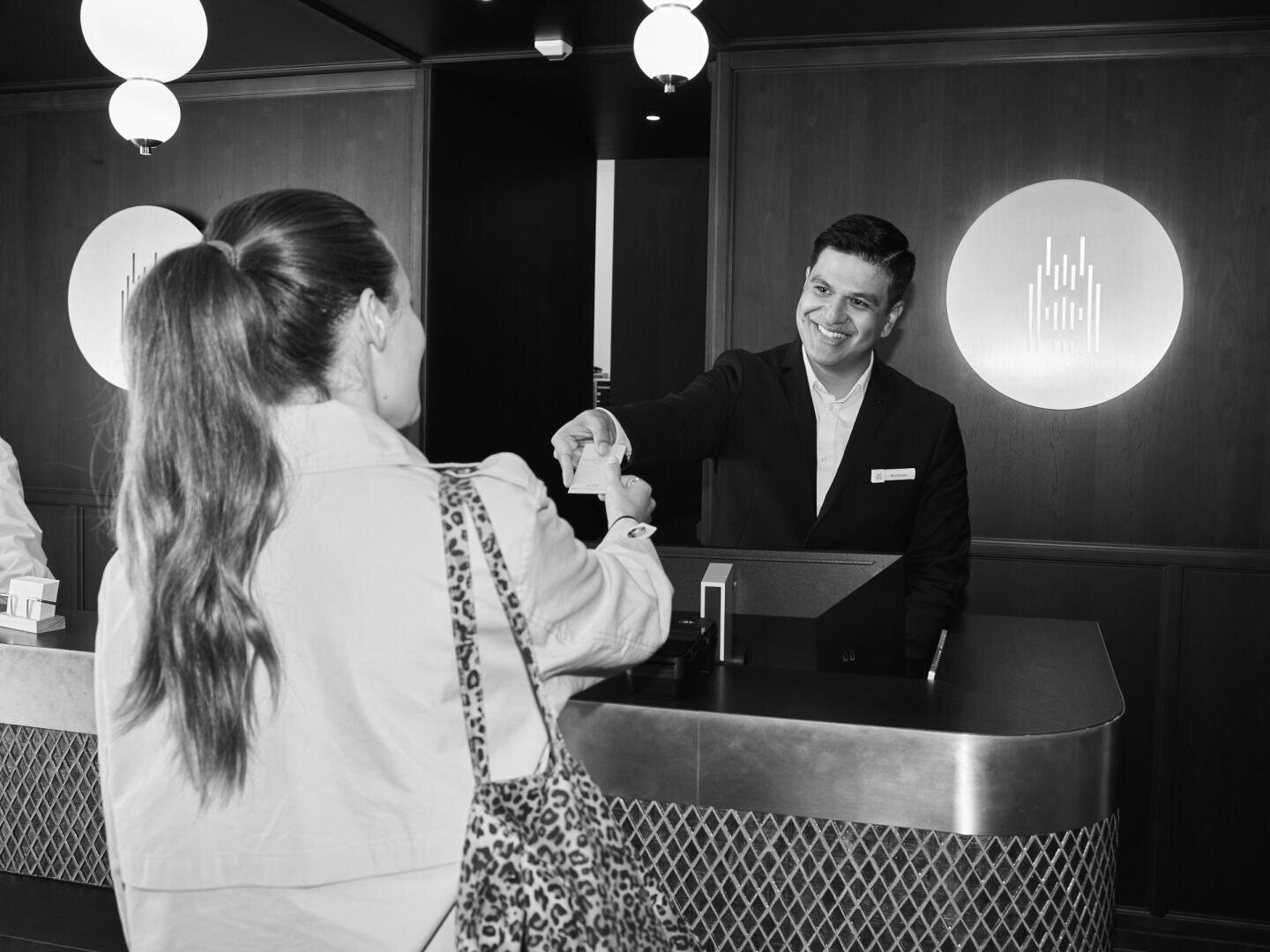 Ein Hotelangestellter an der Rezeption lächelt, als er einem Gast, der am Check-in-Schalter steht, eine Schlüsselkarte überreicht. Der Gast steht mit dem Rücken zur Kamera, und die Umgebung ist modern und gut beleuchtet.