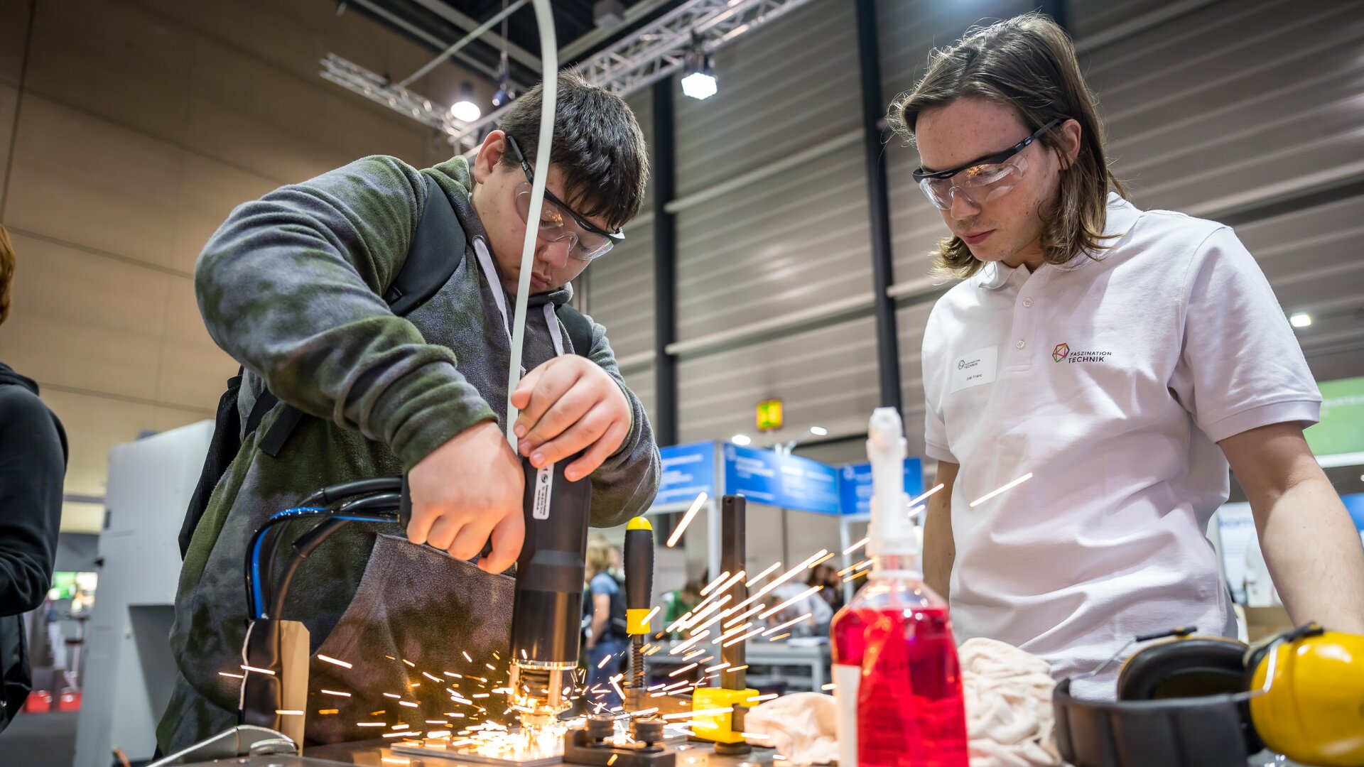 Zwei Personen mit Schutzbrillen arbeiten mit einem Elektrowerkzeug, das Funken auf einer Metalloberfläche erzeugt. Einer bedient das Werkzeug, während der andere beobachtet. Die Sicherheitsausrüstung und die Flaschen stehen auf dem Tisch in einer industriellen Umgebung.