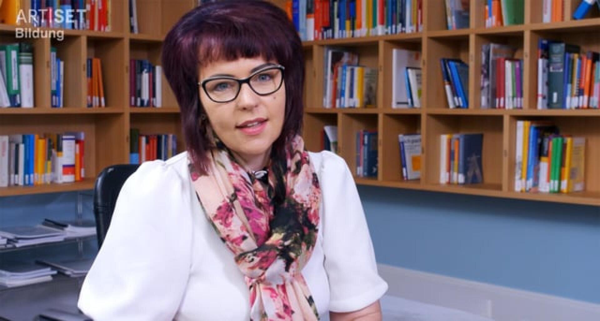Eine Frau mit kurzen dunklen Haaren und Brille, die ein weißes Oberteil und einen geblümten Schal trägt, sitzt vor Bücherregalen voller Bücher in einem hell beleuchteten Büro oder einer Bibliothek.