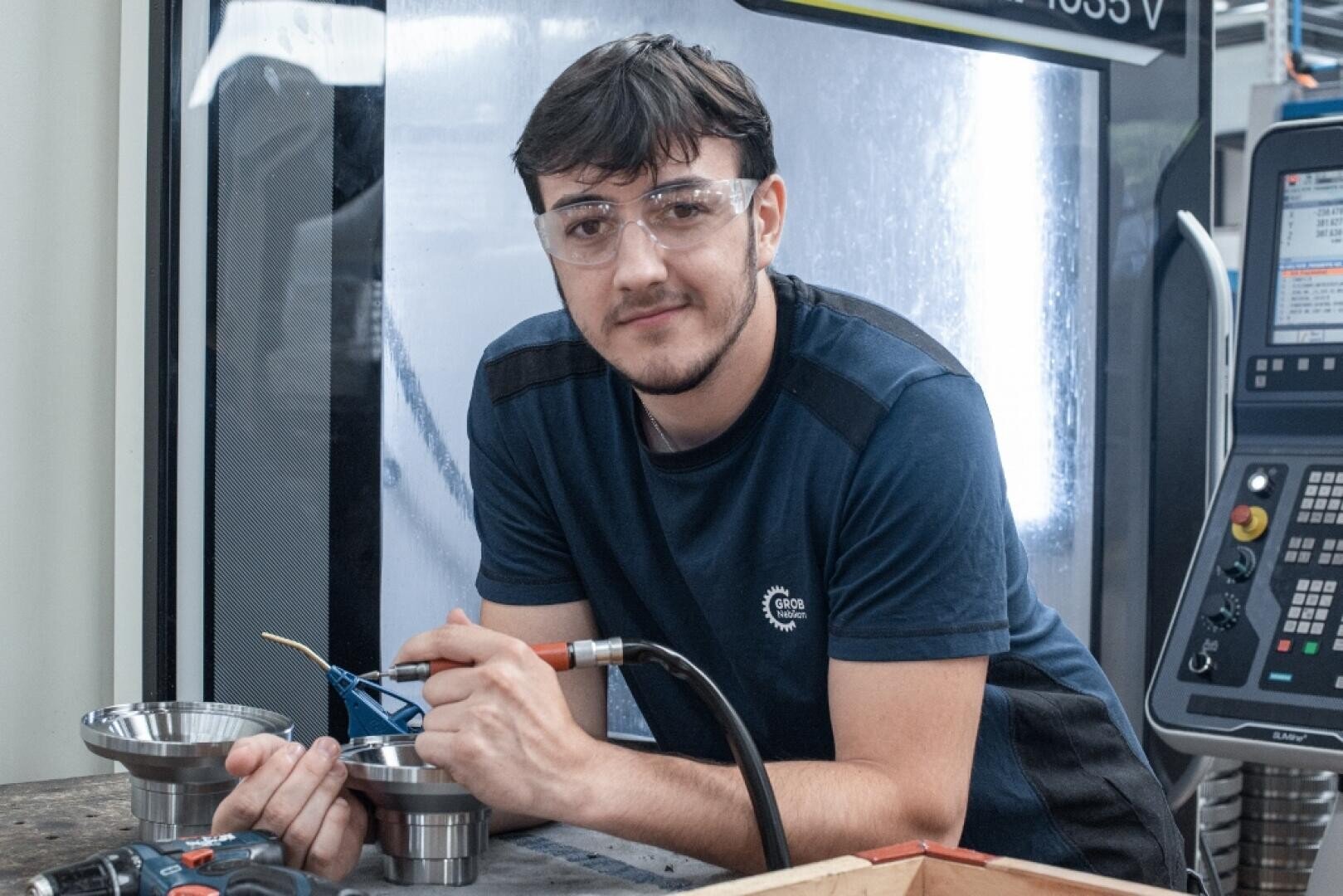 Ein junger Mann mit Schutzbrille lächelt, während er ein Werkzeug an einem Arbeitsplatz mit Metallteilen neben sich hält. Er befindet sich in einer industriellen Umgebung mit Maschinen und einer Schalttafel im Hintergrund.
