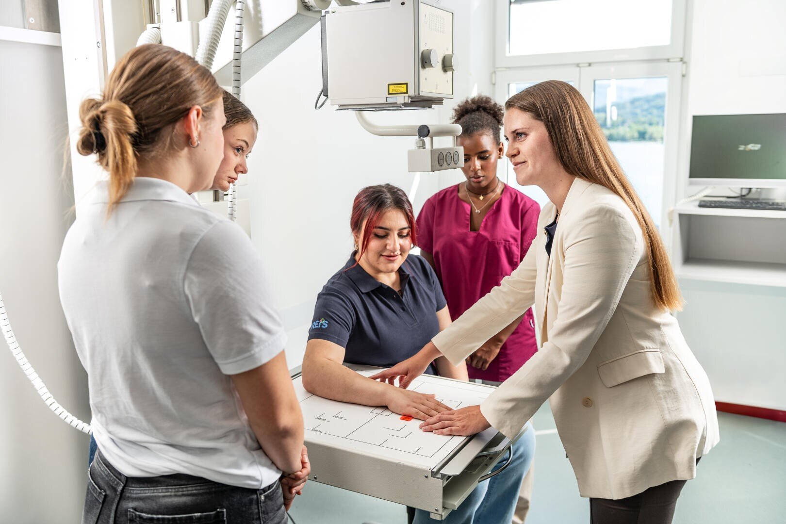 Vier Frauen stehen um eine andere Frau herum, die mit ihrem Arm auf einem Röntgengerät sitzt, während eine Person in einem weißen Blazer die Gruppe in einem hellen medizinischen Raum anleitet.