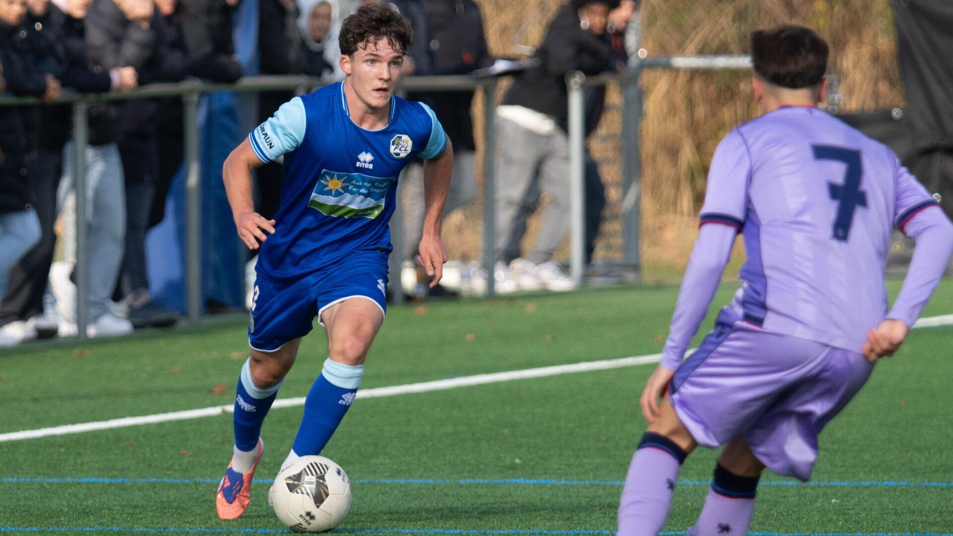 Ein Fußballspieler in einem blauen Trikot dribbelt den Ball, während er einem Gegner in einem violetten Trikot auf einem Außenplatz gegenübersteht; im Hintergrund beobachten Zuschauer das Geschehen.