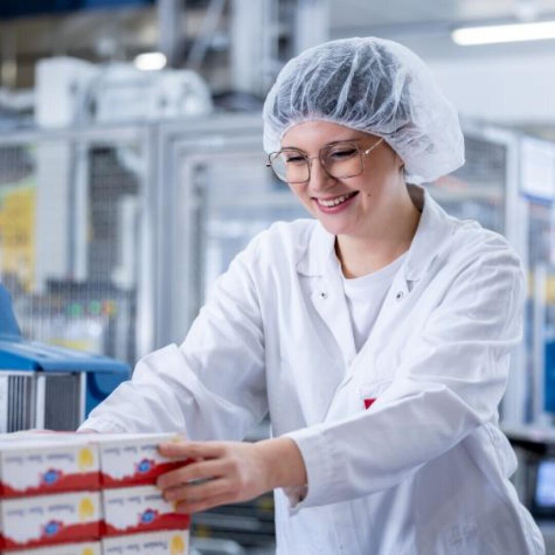 Ein lächelnder Arbeiter in weißem Laborkittel, Haarnetz und Brille stapelt Kisten in einer Lebensmittelverarbeitungsanlage. Im Hintergrund sind Industriemaschinen und eine saubere, organisierte Umgebung zu sehen.