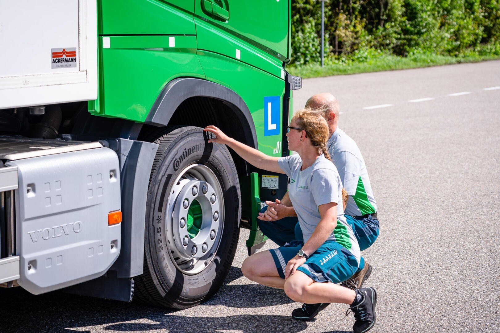 Zwei Menschen in Uniform hocken neben einem grünen Lkw und untersuchen an einem sonnigen Tag aufmerksam den Vorderreifen. Der Lkw steht auf einer asphaltierten Straße, im Hintergrund ist Grün zu sehen.