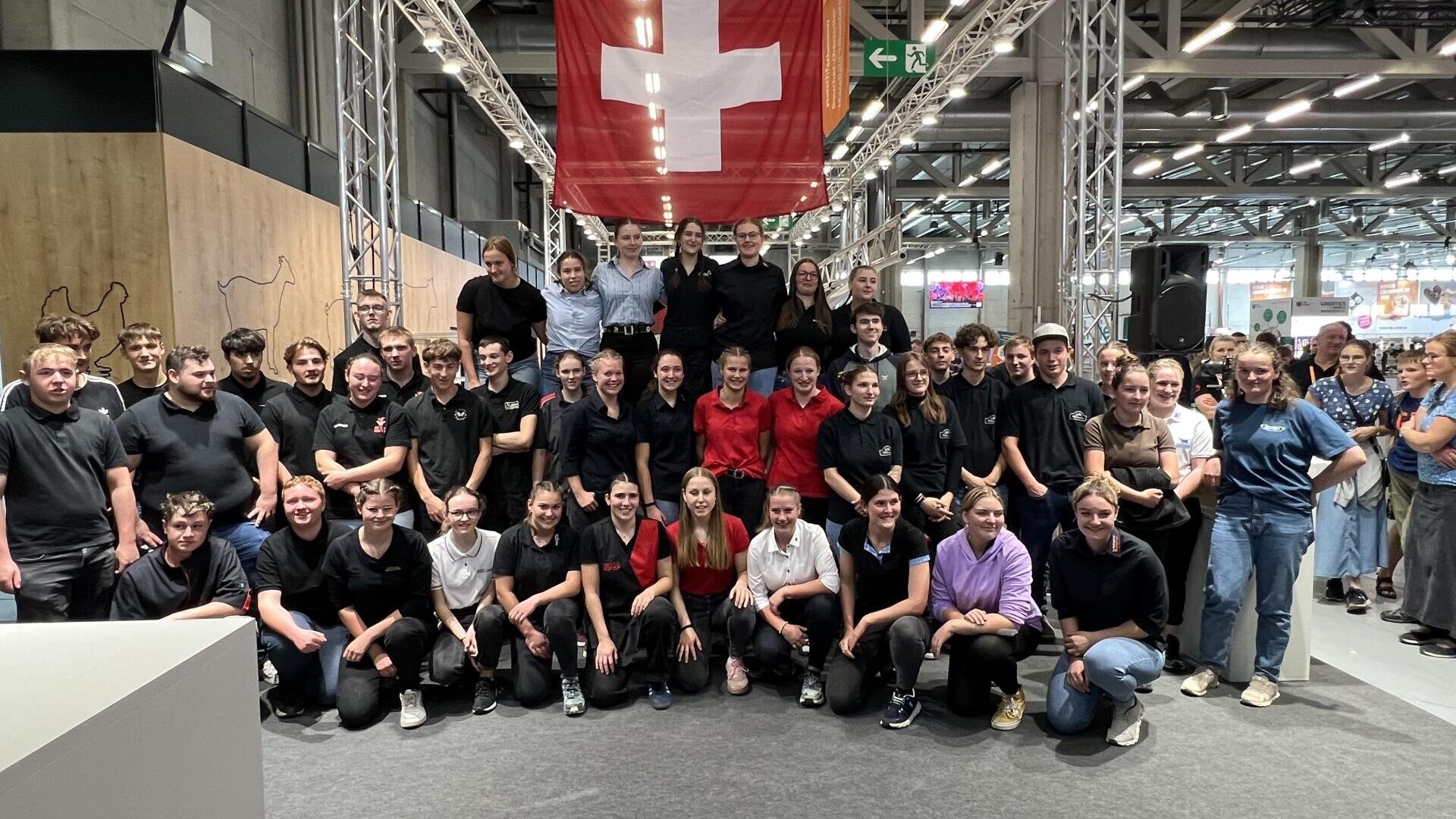Eine große Gruppe von Menschen posiert gemeinsam in einem Innenraum unter einer hängenden Schweizer Flagge. Die meisten tragen schwarze oder rote Uniformen, während einige wenige auf der rechten Seite Freizeitkleidung tragen. Der Schauplatz scheint eine öffentliche Veranstaltung oder Ausstellung zu sein.