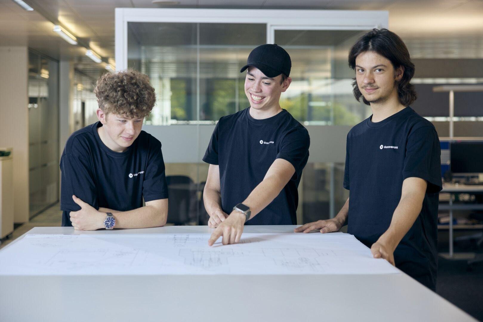 Drei junge Männer in passenden schwarzen T-Shirts stehen in einem modernen Büro um einen Tisch herum und prüfen und diskutieren gemeinsam architektonische Entwürfe. Einer zeigt auf die Pläne, alle scheinen beschäftigt zu sein.