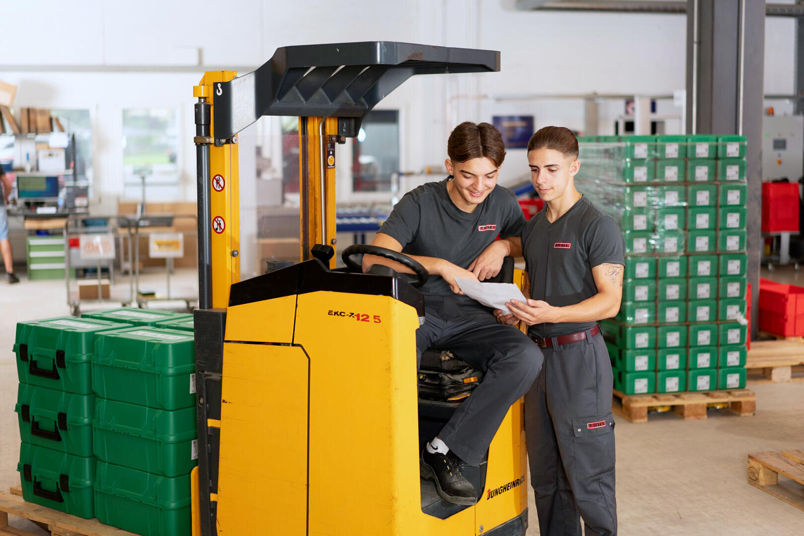Zwei Lagerarbeiter in grauen Uniformen, die neben einem gelben Gabelstapler stehen und ein Dokument durchsehen.