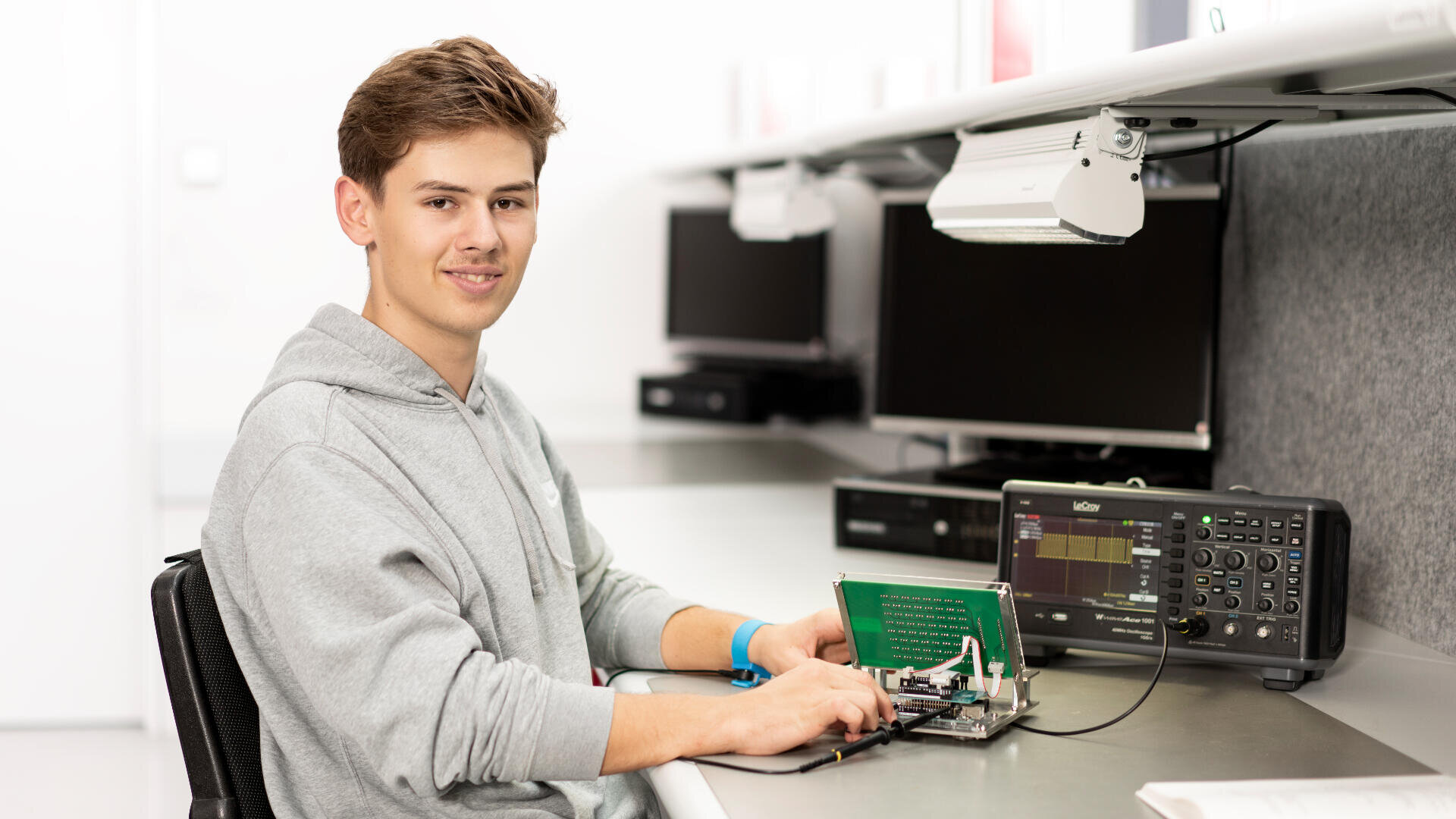 Ein junger Mann im grauen Kapuzenpulli sitzt an einem Schreibtisch und arbeitet mit Messgeräten und Monitoren in einer hellen, modernen Laborumgebung an einer elektronischen Platine.
