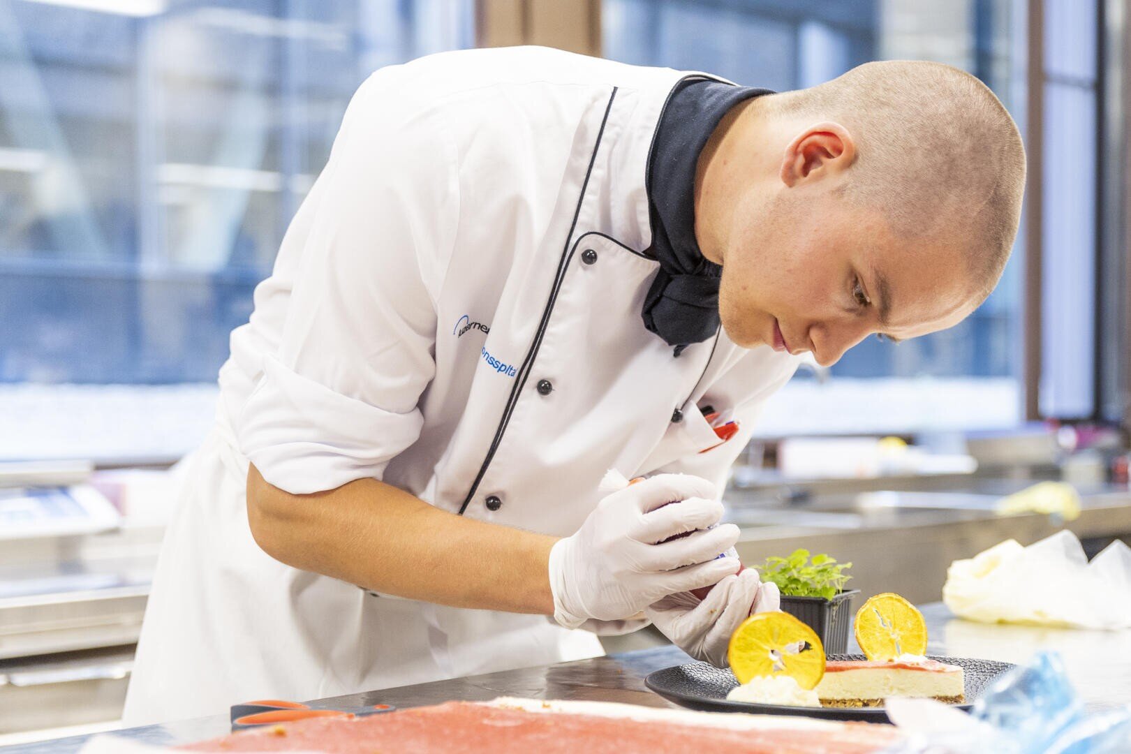 Ein Koch in weißer Uniform und Handschuhen dekoriert sorgfältig einen Teller mit Orangenscheiben und Garnierungen in einer modernen Küche.