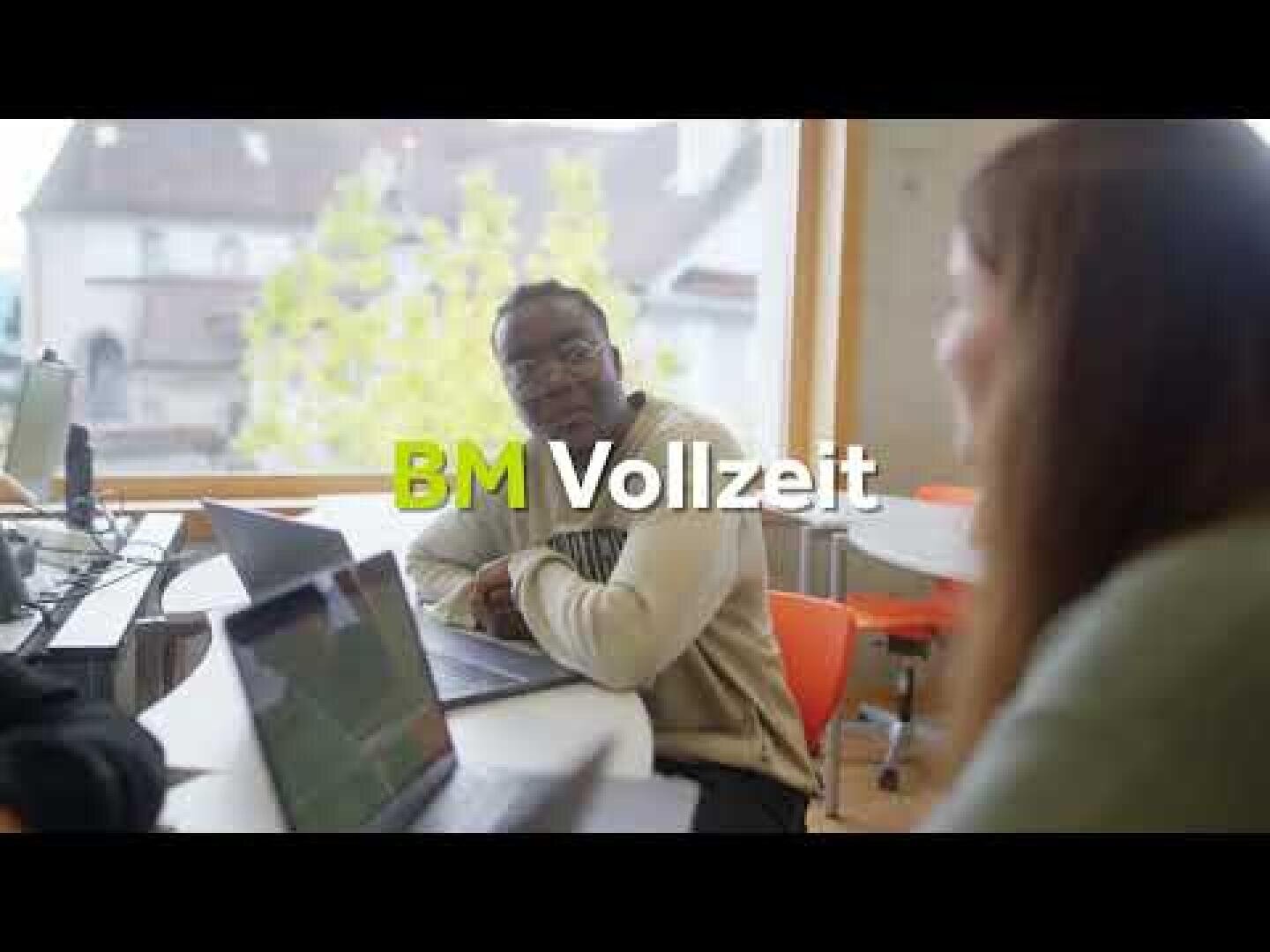 Zwei Personen sitzen mit ihren Laptops an einem Tisch in einem hellen Raum. In der Bildmitte ist der Schriftzug „BM Vollzeit“ zu sehen. Durch ein großes Fenster im Hintergrund sind Bäume und Gebäude zu erkennen.