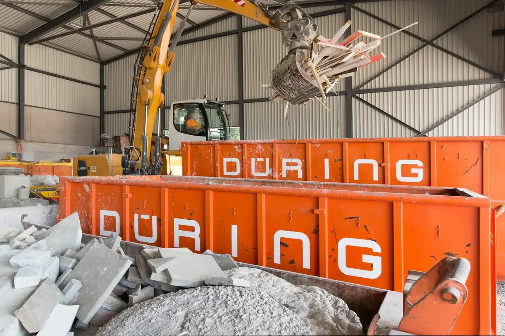 Ein Bagger kippt Bauschutt in einen orangefarbenen Müllcontainer mit der Aufschrift DÜRING in einer Lagerhalle, in der sich Stapel von Betonbruch und anderen Baumaterialien befinden.
