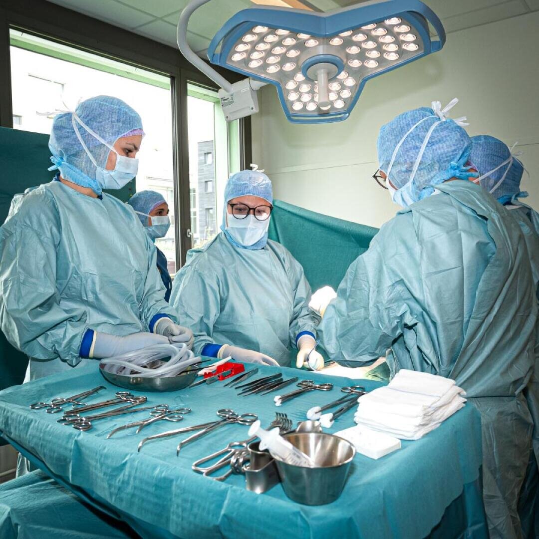 Vier Chirurgen in blauen OP-Kitteln und Masken operieren in einem hell beleuchteten Raum. Im Vordergrund liegen die chirurgischen Instrumente ordentlich auf einem grünen Tisch. Über ihnen hängt eine große Operationsleuchte.