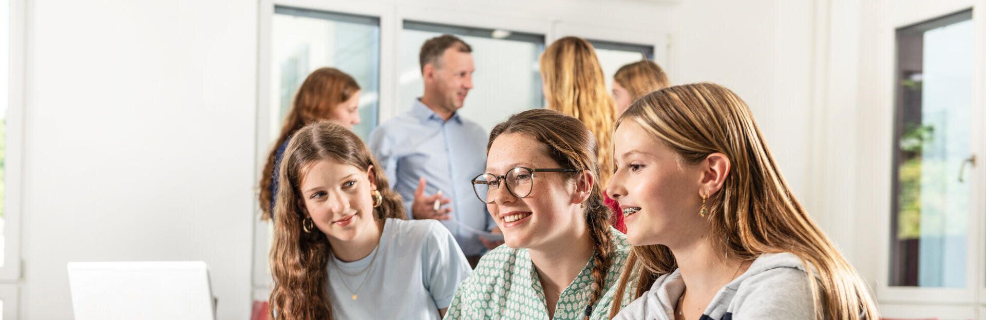 Drei Mädchen im Teenageralter lächeln und arbeiten gemeinsam an einem Tisch mit einem Laptop. Im Hintergrund unterhalten sich Erwachsene und andere Schüler in einem hellen, modernen Klassenzimmer.