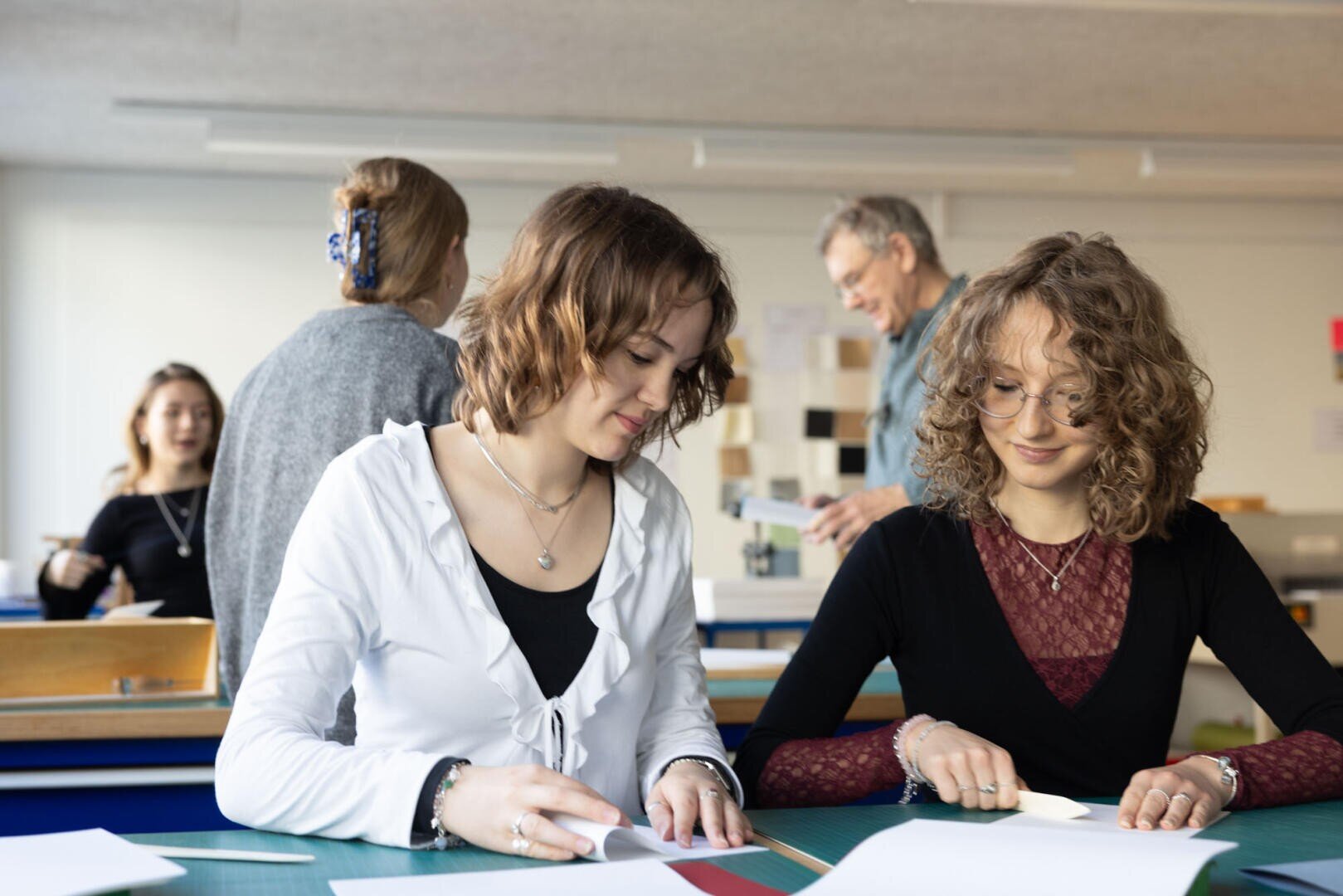 Zwei junge Frauen sitzen an einem Tisch und arbeiten an Papieren, während drei andere Personen, darunter ein älterer Mann, im Hintergrund eines hellen Klassenzimmers in Gespräche und Aktivitäten vertieft sind.
