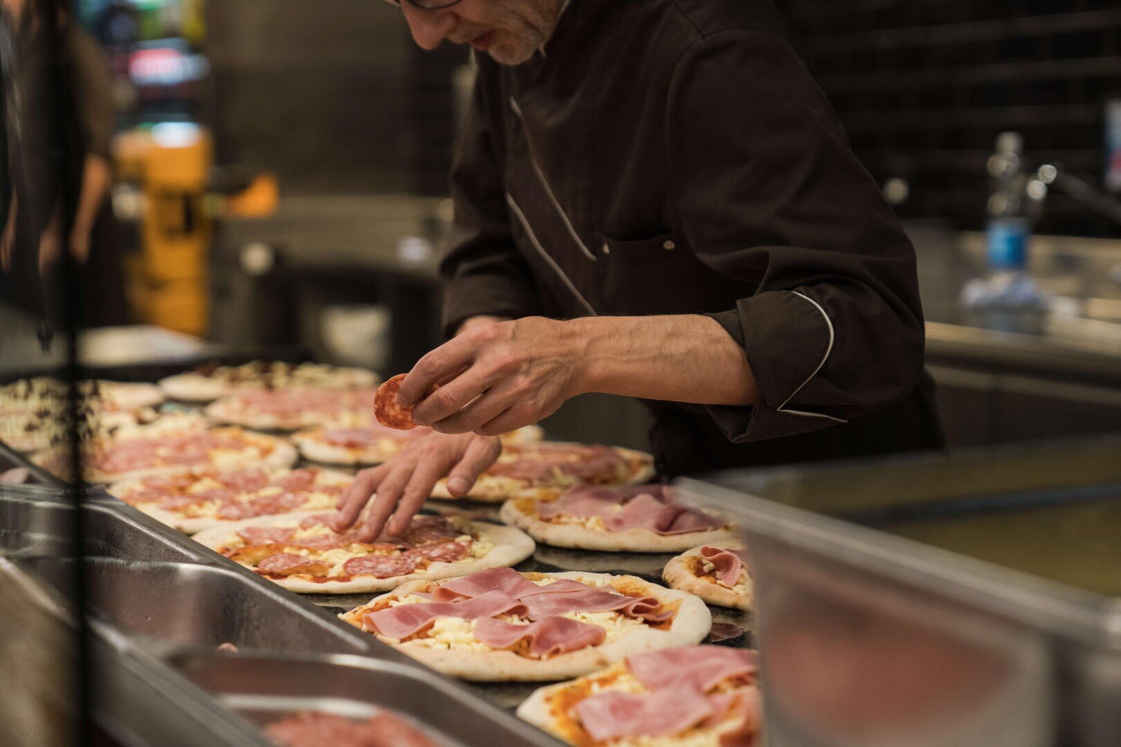 Ein Koch in dunkler Uniform bereitet mehrere rohe Pizzen zu und belegt den Teig und den Belag auf einem Tresen in einer Restaurantküche sorgfältig mit Fleischscheiben.