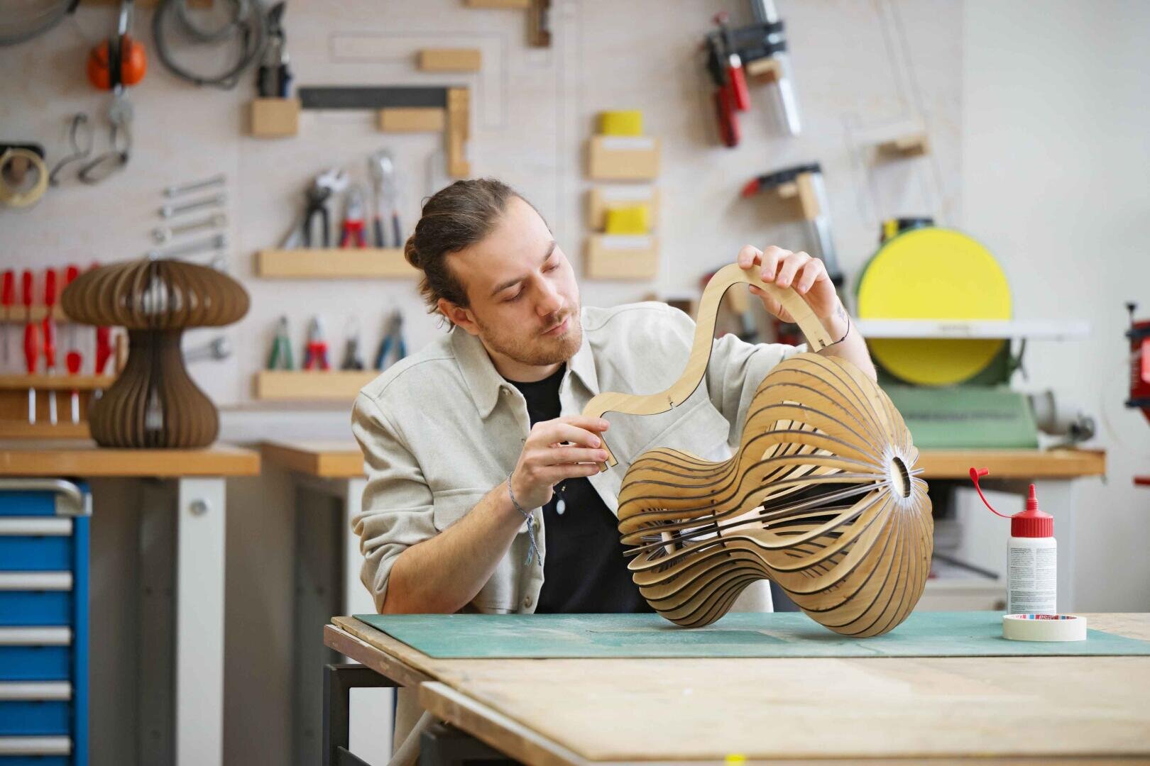 Ein Mann baut eine hölzerne, skulpturale Lampe in einer Werkstatt zusammen, umgeben von Werkzeugen und Materialien. Im Hintergrund sind eine weitere ähnliche Lampe und Arbeitsmaterialien zu sehen.