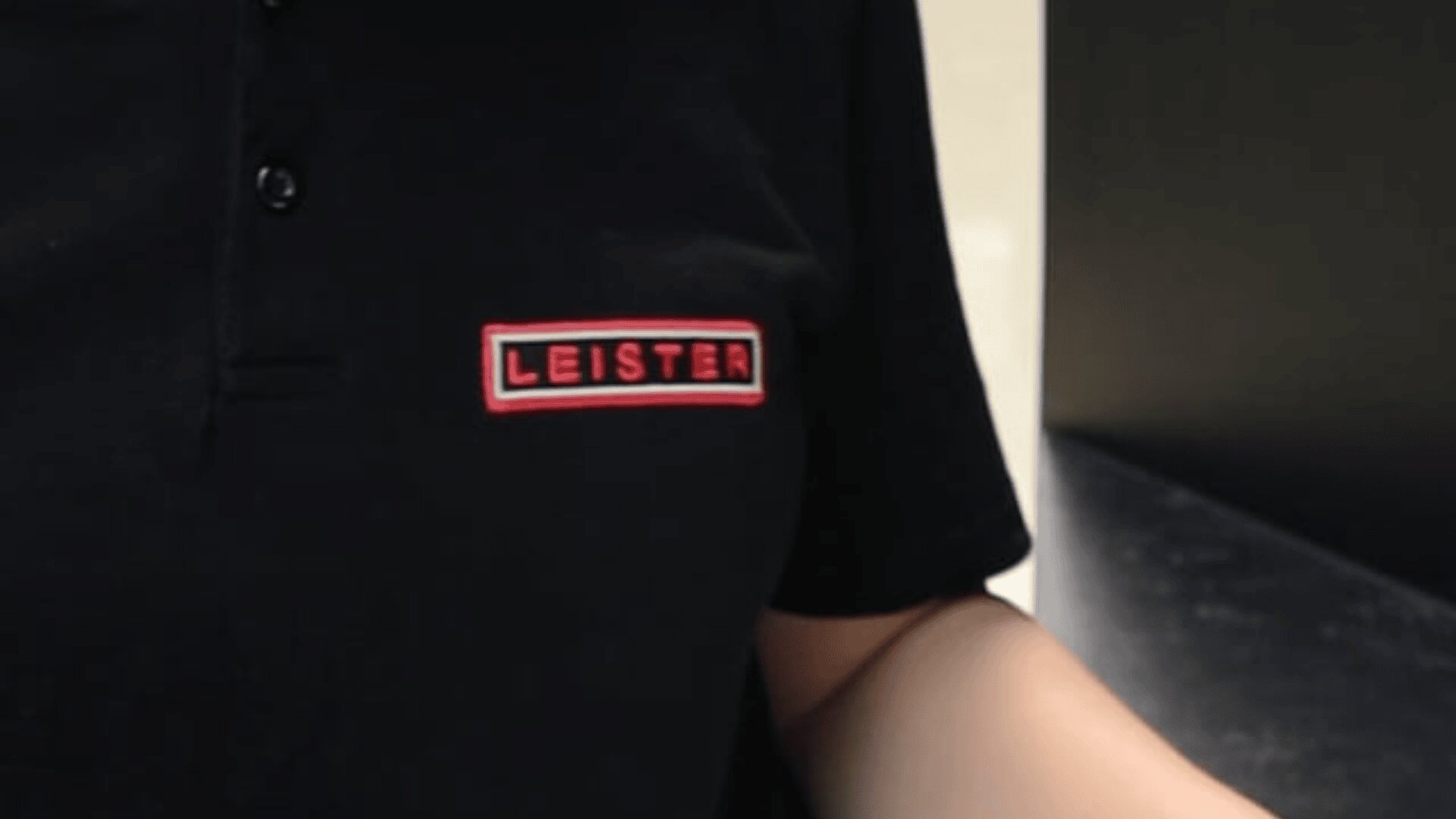 Eine Person, die ein schwarzes Hemd mit Kragen und einem rot-weißen rechteckigen LEISTER-Logoaufnäher auf der linken Brustseite trägt.