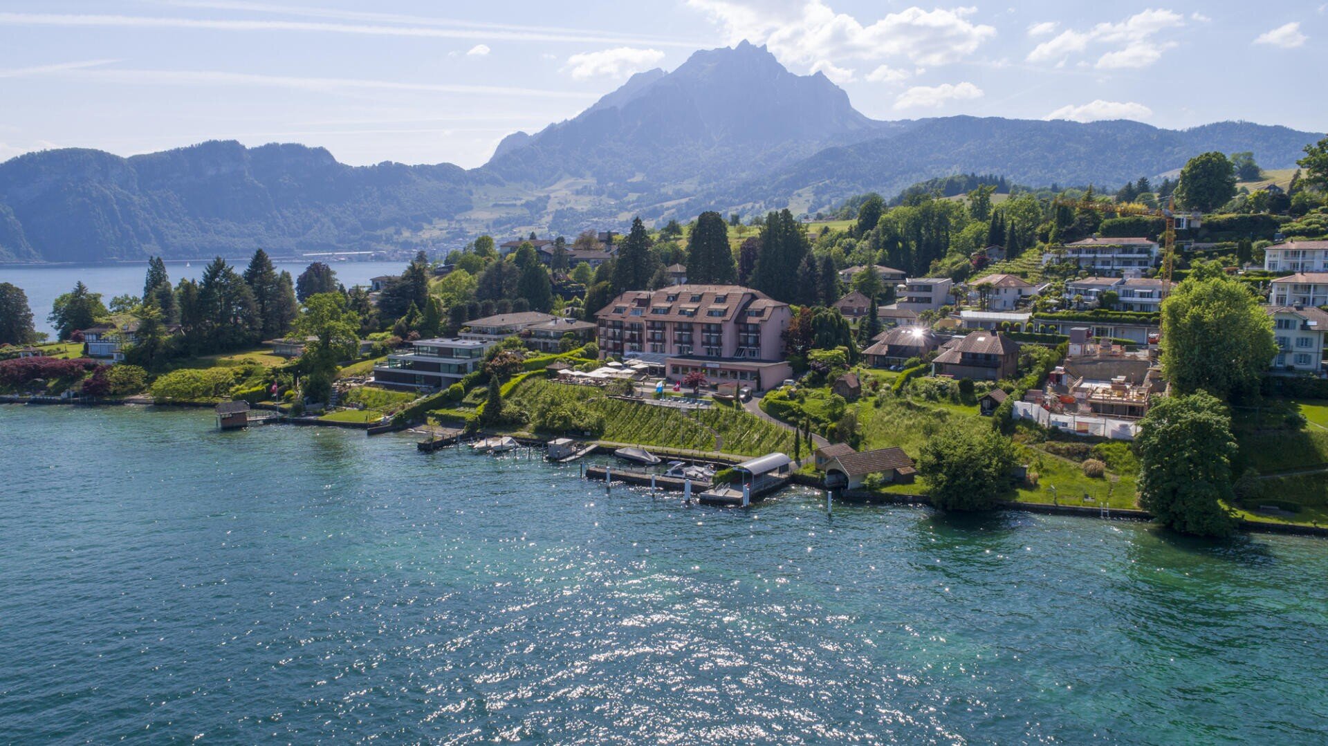Luftaufnahme von Häusern und Gebäuden am See, eingebettet in üppiges Grün, mit einem tiefblauen See im Vordergrund und hoch aufragenden Bergen unter einem teilweise bewölkten Himmel im Hintergrund.