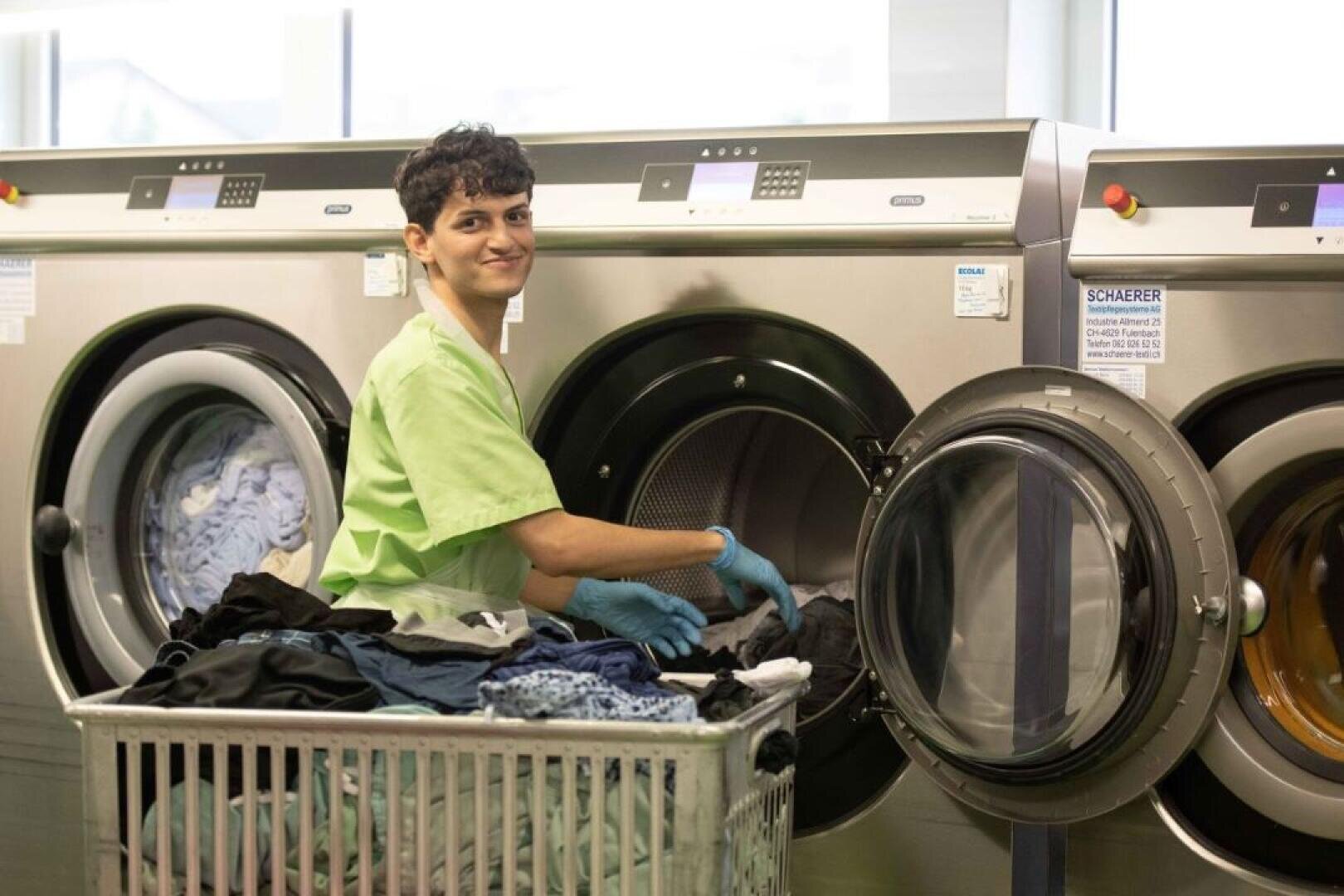 Eine Person, die ein grünes Hemd und blaue Handschuhe trägt, lächelt, während sie in einem Waschsalon Wäsche aus einem Wäschewagen in eine Industriewaschmaschine lädt.