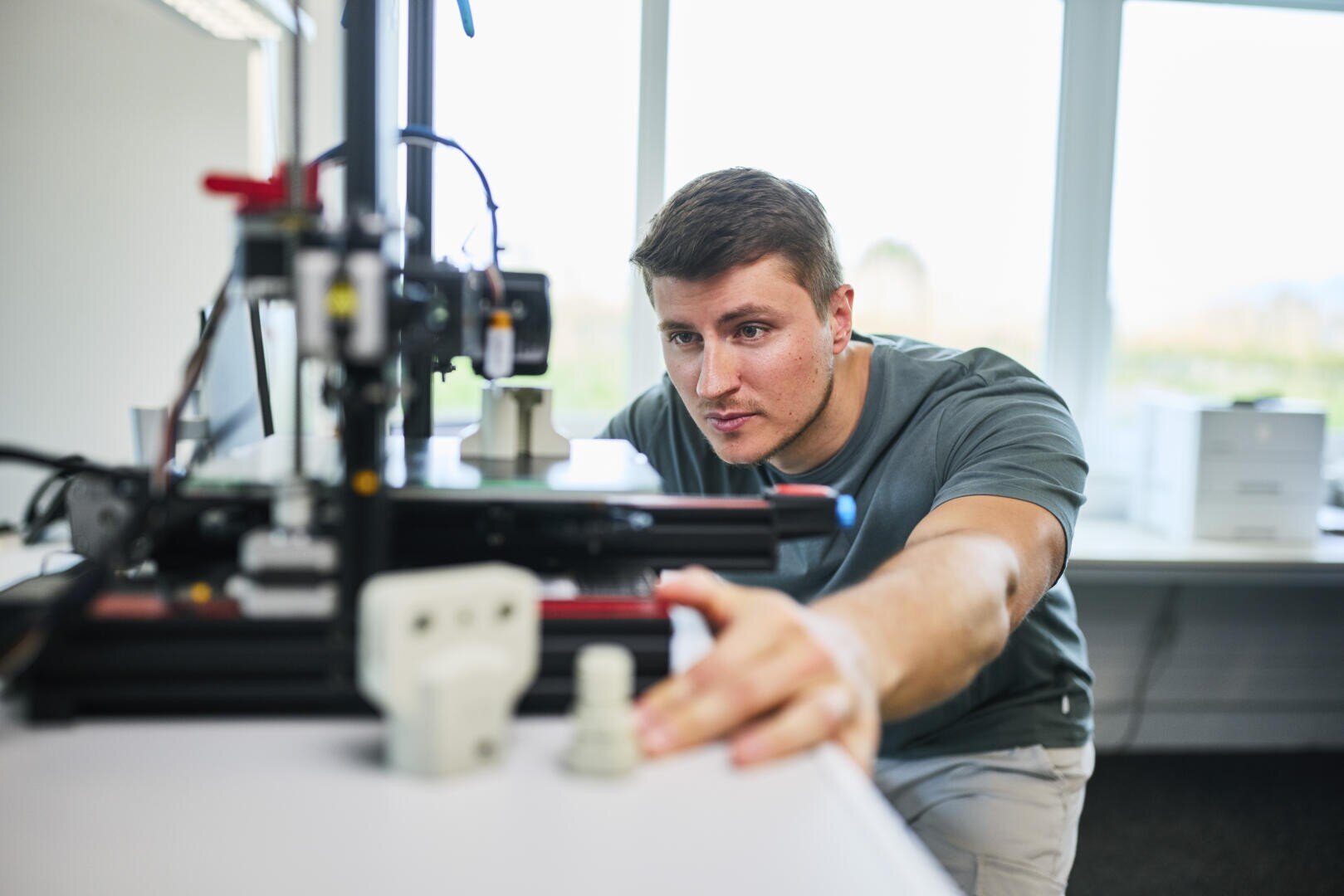 Ein Mann konzentriert sich, während er einen 3D-Drucker auf einem Schreibtisch einstellt, mit verschiedenen kleinen gedruckten Objekten im Vordergrund, in einer hellen Büroumgebung mit großen Fenstern.
