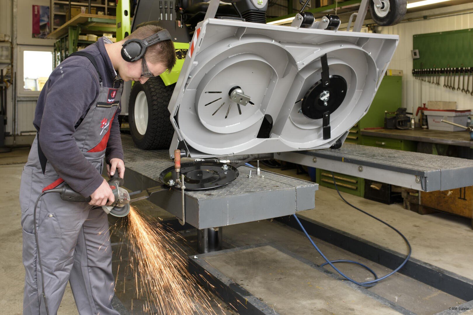 Eine Person mit Gehörschutz benutzt einen Winkelschleifer und erzeugt dabei Funken, während sie in einer Werkstatt auf dem erhöhten Deck eines Rasenmähers arbeitet. Im Hintergrund sind Werkzeuge und Geräte zu sehen.