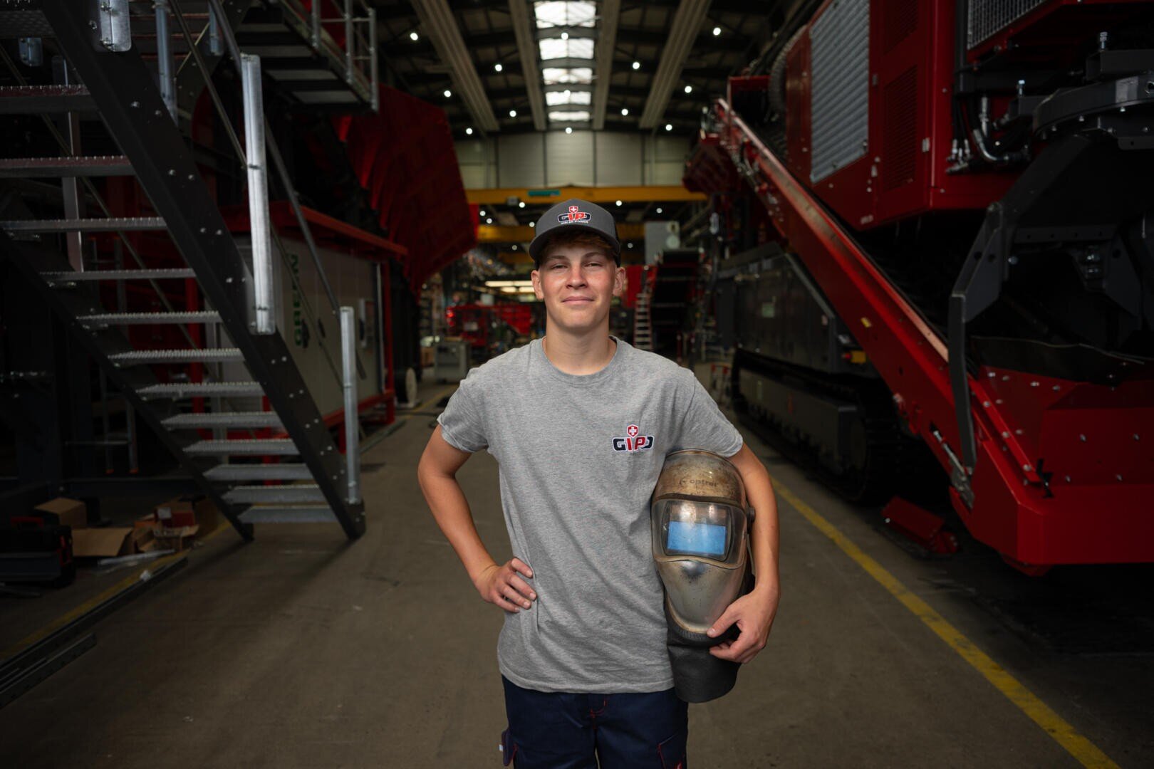 Ein junger Mann in einem grauen T-Shirt und einer Mütze steht in einer Industriehalle, lächelt und hält einen Schweißhelm unter dem Arm. Im Hintergrund sind große rote Maschinen zu sehen.
