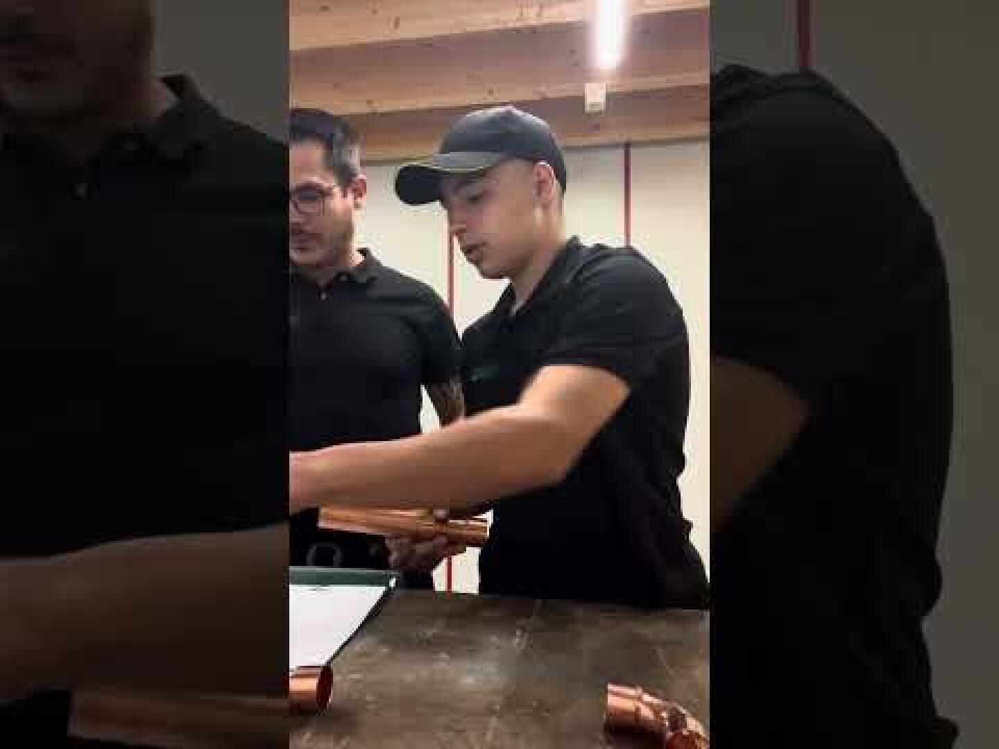 Zwei Männer in schwarzen Hemden stehen an einem Arbeitstisch und hantieren mit Kupferrohren, wobei ein Mann eine schwarze Mütze trägt. Sie scheinen gemeinsam in einem Innenraum unter hölzernen Deckenbalken zu arbeiten.