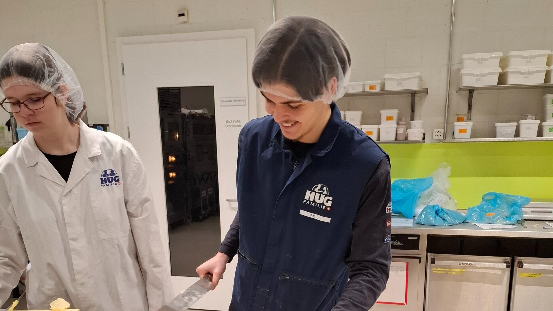 Zwei Personen mit Haarnetzen und Uniformen arbeiten in einer Küche oder einem Labor. Die Person auf der rechten Seite lächelt, während sie ein Werkzeug in der Hand hält. Im Hintergrund sind Regale mit Behältern und ein Kühlschrank zu sehen. Auf beiden Uniformen ist das HUG-Logo zu sehen.