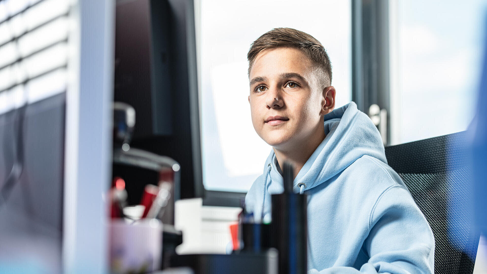 Ein Jugendlicher in einem hellblauen Kapuzenpulli sitzt an einem Schreibtisch und schaut aufmerksam auf einen Computerbildschirm in einem hellen, modernen Raum mit Büromaterial im Vordergrund.