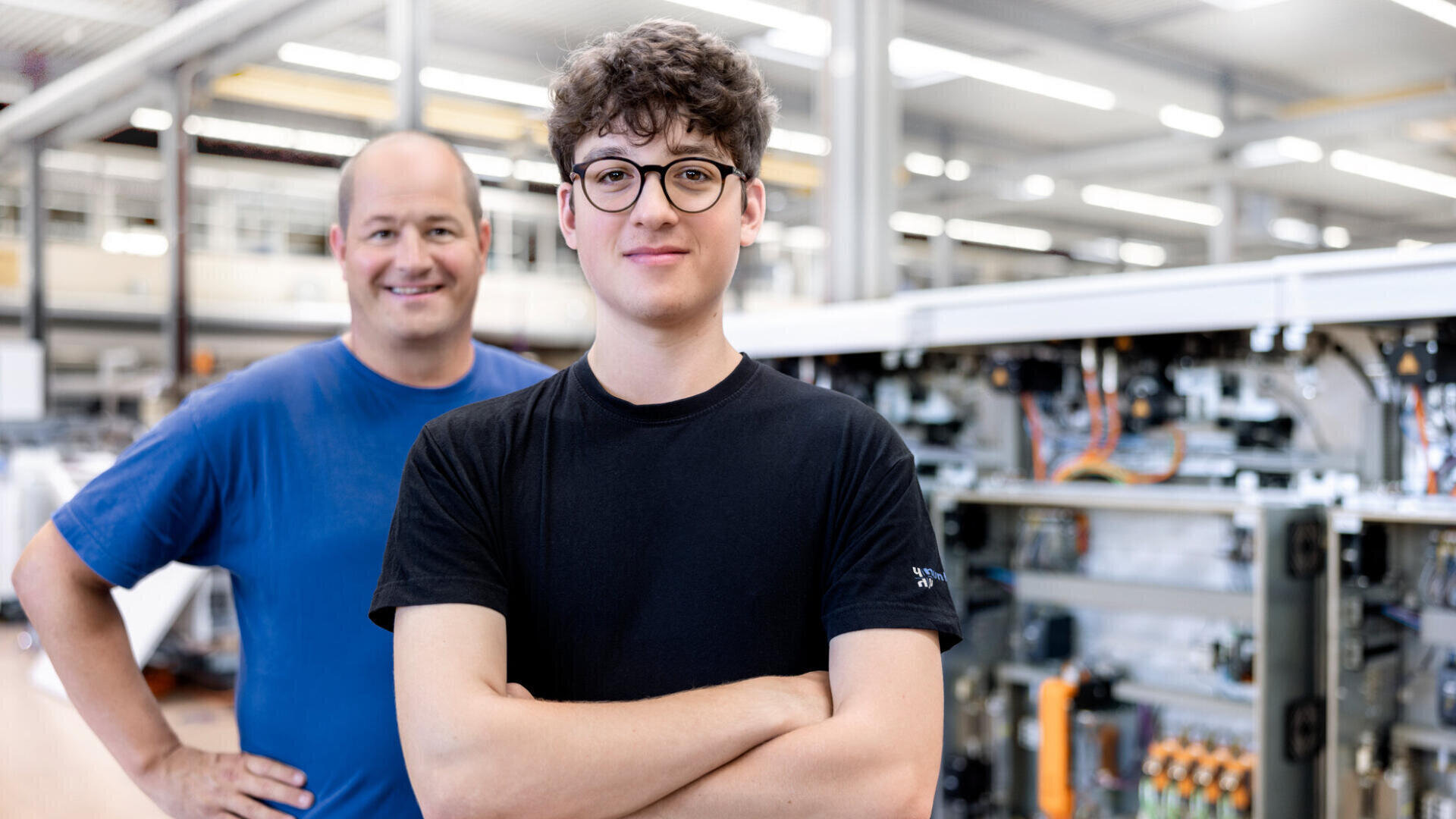 Zwei Personen stehen in einer modernen Industrieumgebung. Eine junge Person mit lockigem Haar und Brille steht selbstbewusst im Vordergrund, die Arme verschränkt, während eine ältere Person in einem blauen Hemd im Hintergrund in der Nähe von Maschinen lächelt.