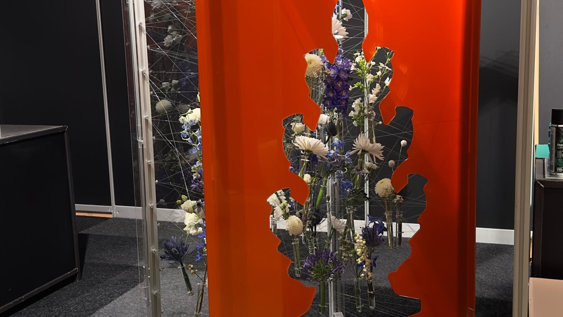Ein Blumenarrangement mit weißen und violetten Blumen wird hinter einer leuchtend orangefarbenen Platte mit einem unregelmäßigen Ausschnitt in einem modernen Innenraum mit schwarzen Wänden und klaren Trennwänden präsentiert.